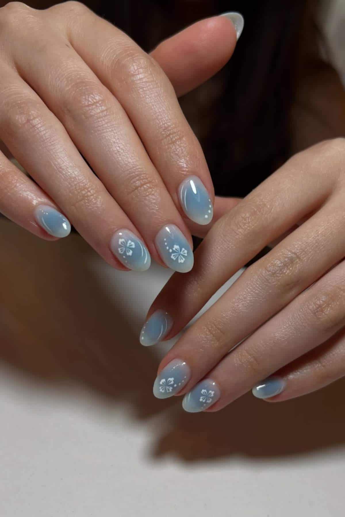 Two hands with almond-shaped nails painted light blue, some featuring small white floral designs and rhinestone accents.