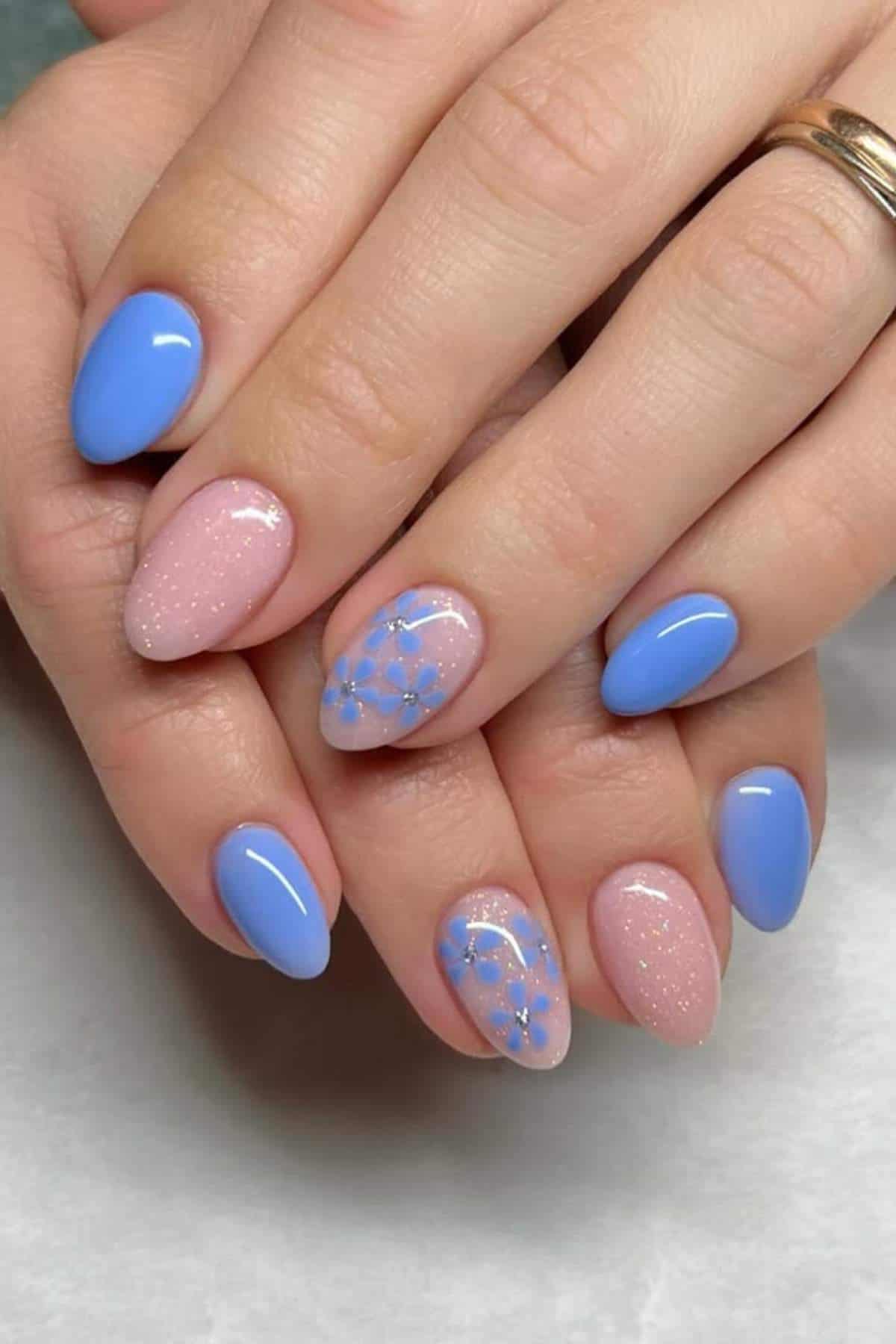 Hands with almond-shaped nails painted blue and pale pink, featuring glitter and blue floral designs on two accent nails.