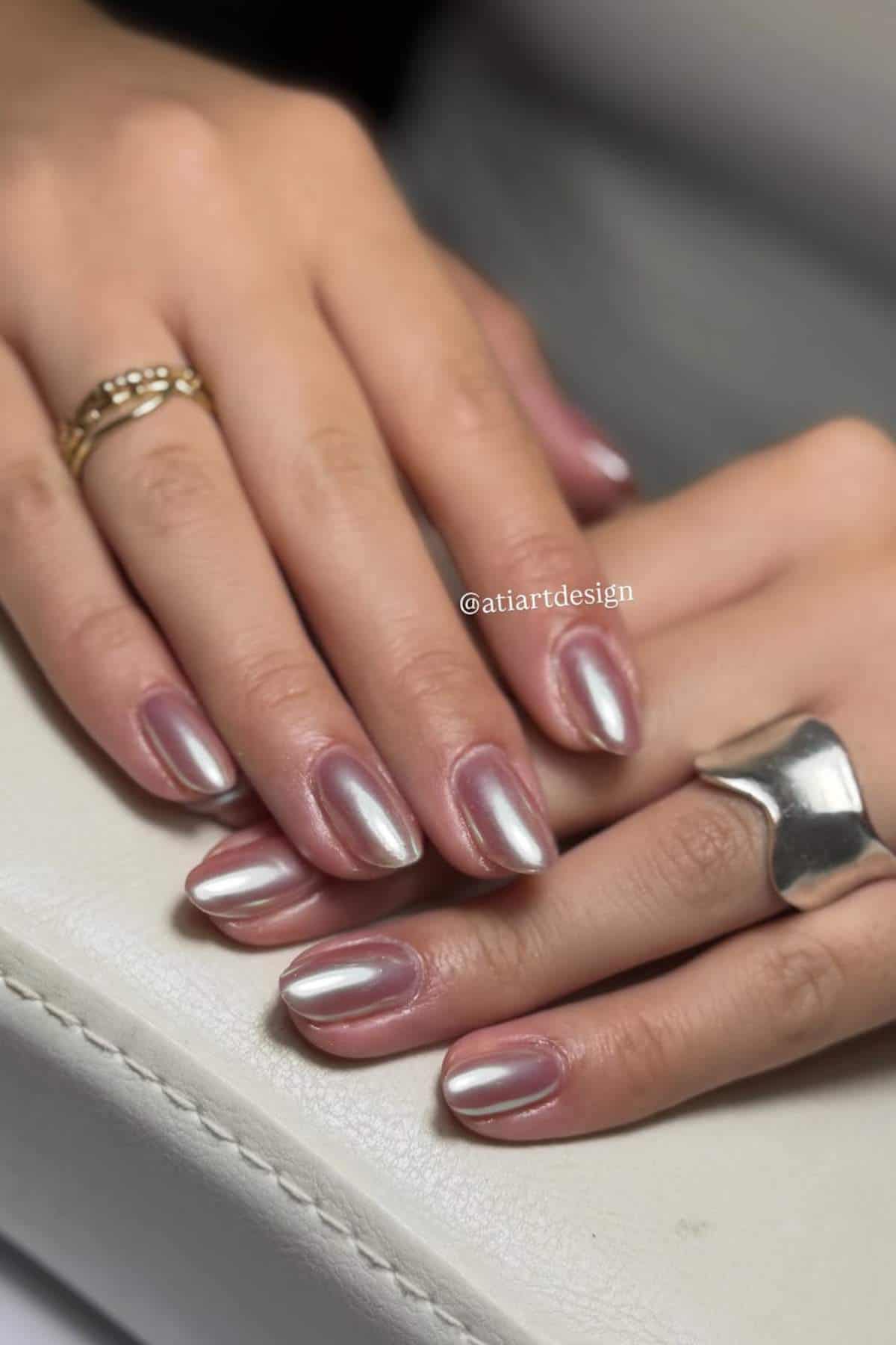 Close-up of hands with short, almond-shaped nails painted in a shiny silver chrome polish, resting on a white surface. The person wears two rings, one gold and one silver.