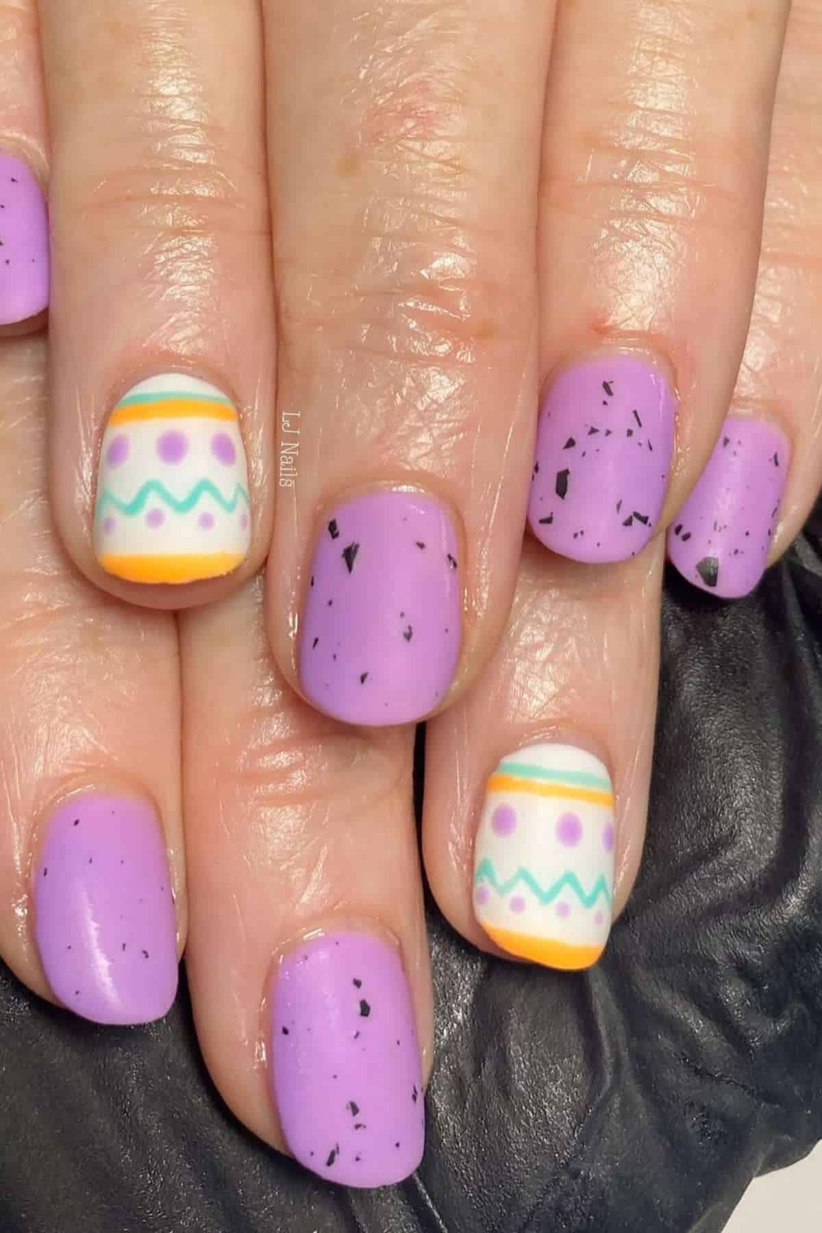 Close-up of hands with pastel purple nails; two nails feature Easter egg-inspired designs with stripes, dots, and zigzags in white, orange, green, and purple.