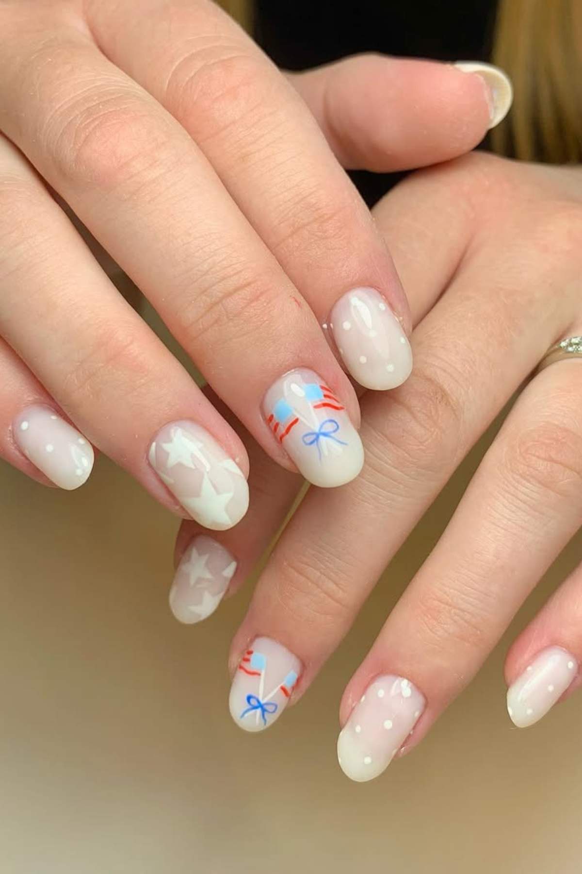 A hand with short, oval nails painted with a pale base color, decorated with white stars, white dots, and red and blue bows.