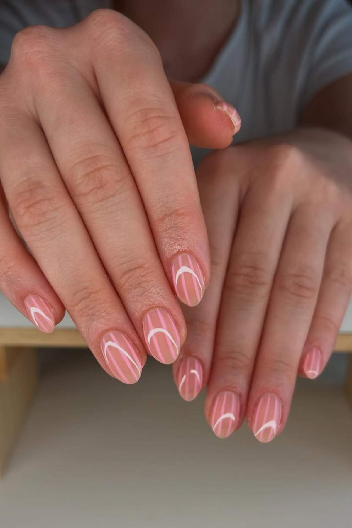Close-up of hands with neatly manicured nails painted in a natural pink shade, featuring a simple white abstract line design on each nail.