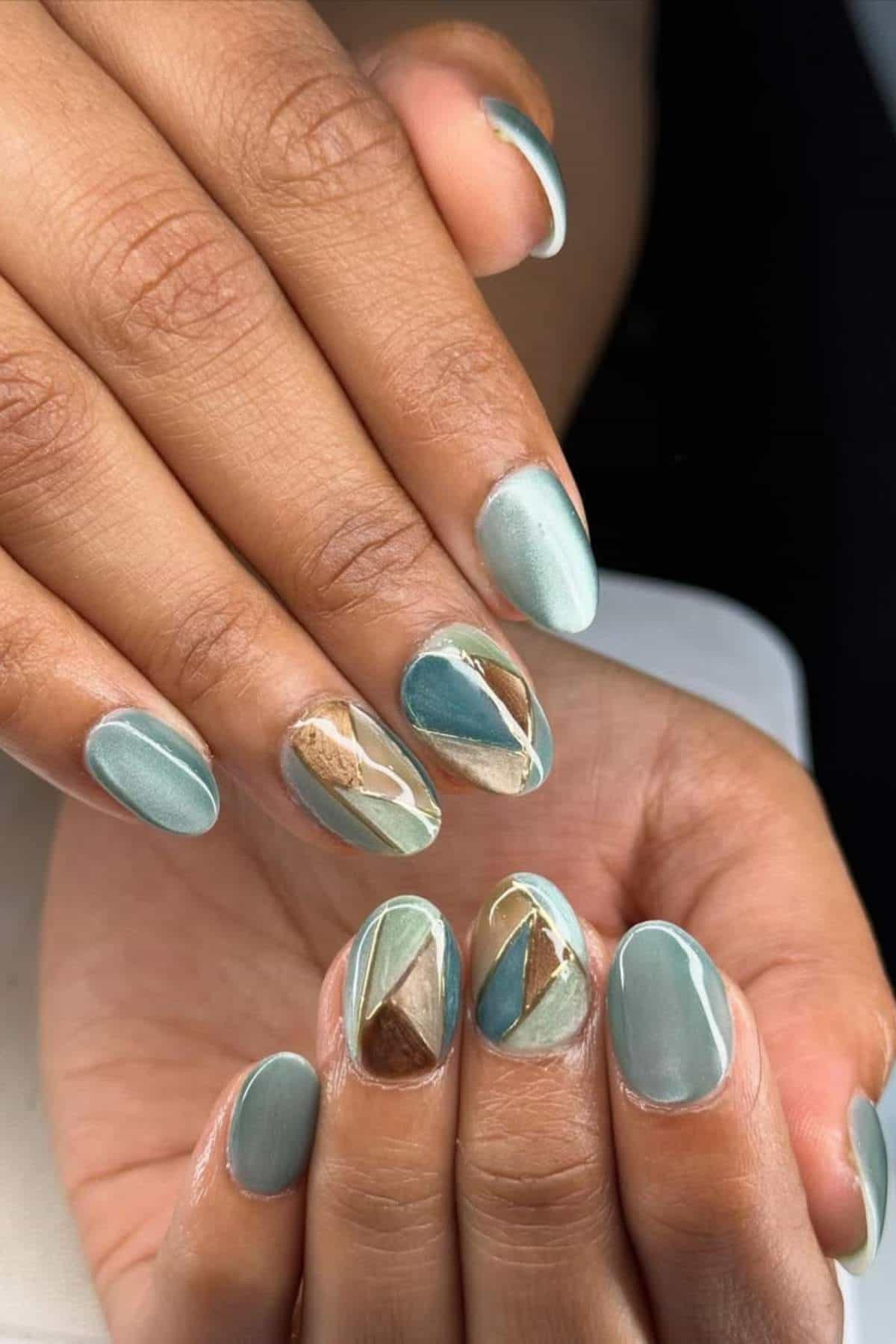 Hands with short oval nails painted in light green polish, featuring geometric designs in gold, brown, and teal on accent nails.