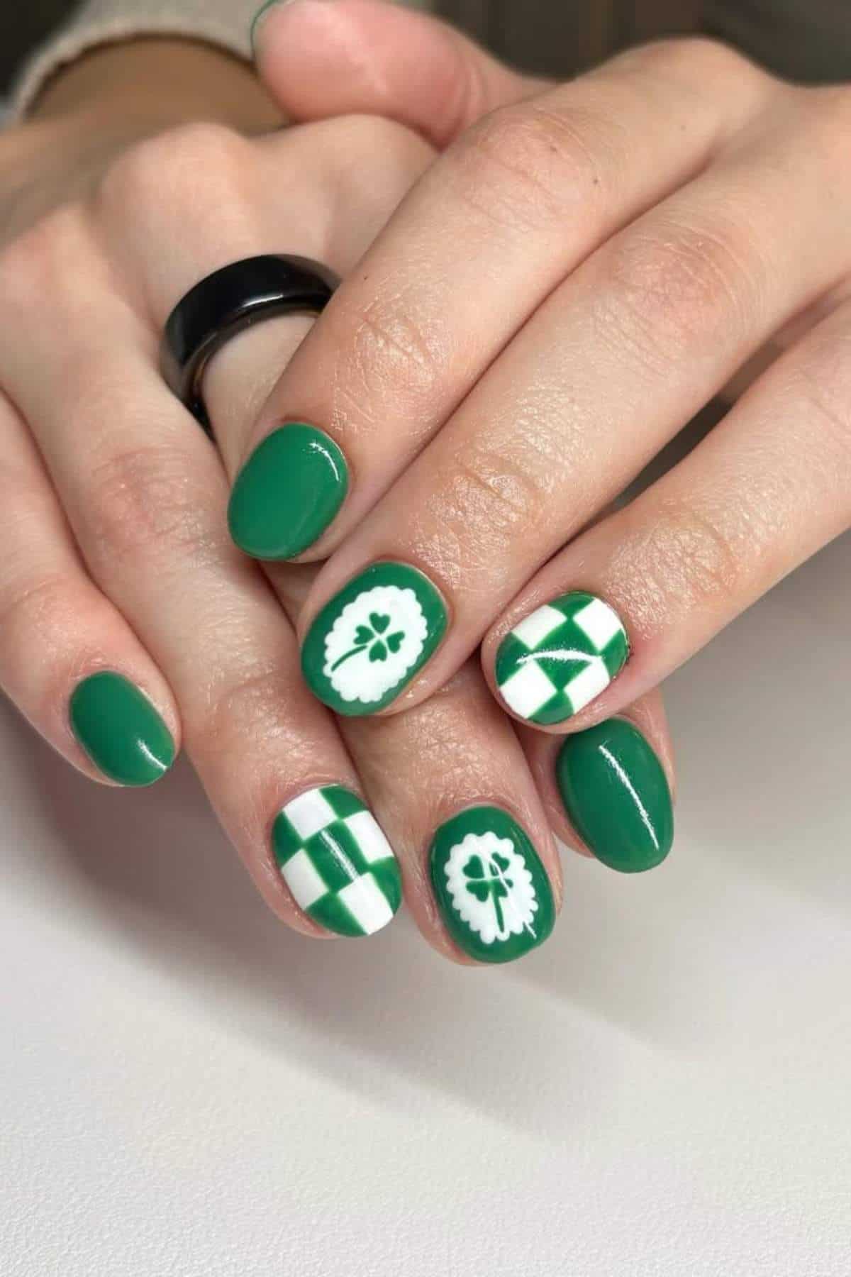 Hands with short, oval-shaped nails painted green and white with plaid and floral patterns on some nails. A black ring is worn on one finger.