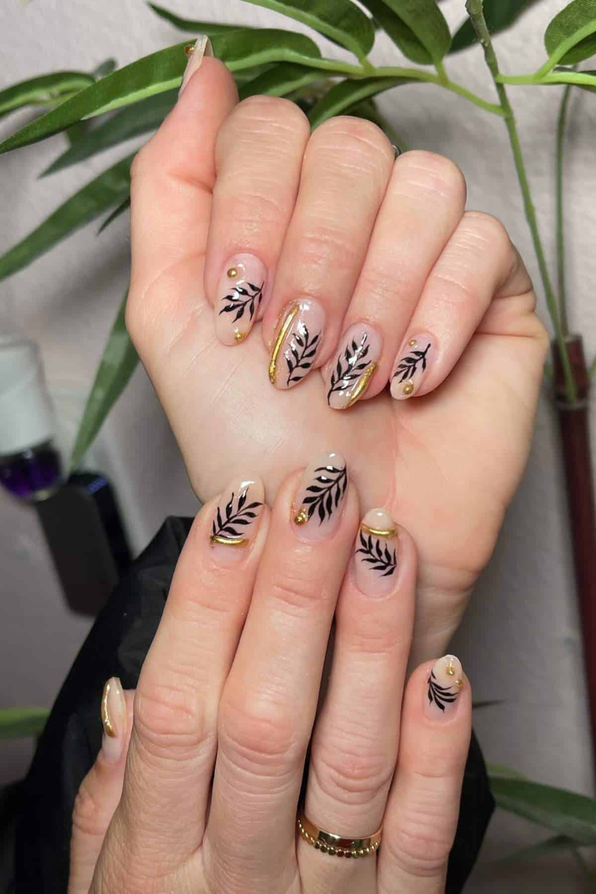 Hands with almond-shaped nails featuring black leaf designs, gold accents, and small gold studs, posed in front of green plant leaves.
