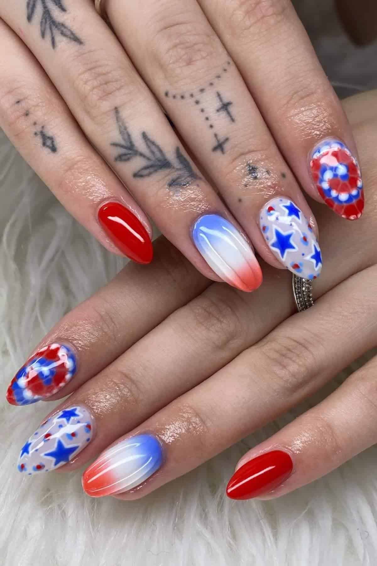 Hands with patriotic-themed manicure featuring red, white, and blue colors, tie-dye patterns, star designs, and ombre effects on long, pointed nails. Tattoos are visible on the fingers.