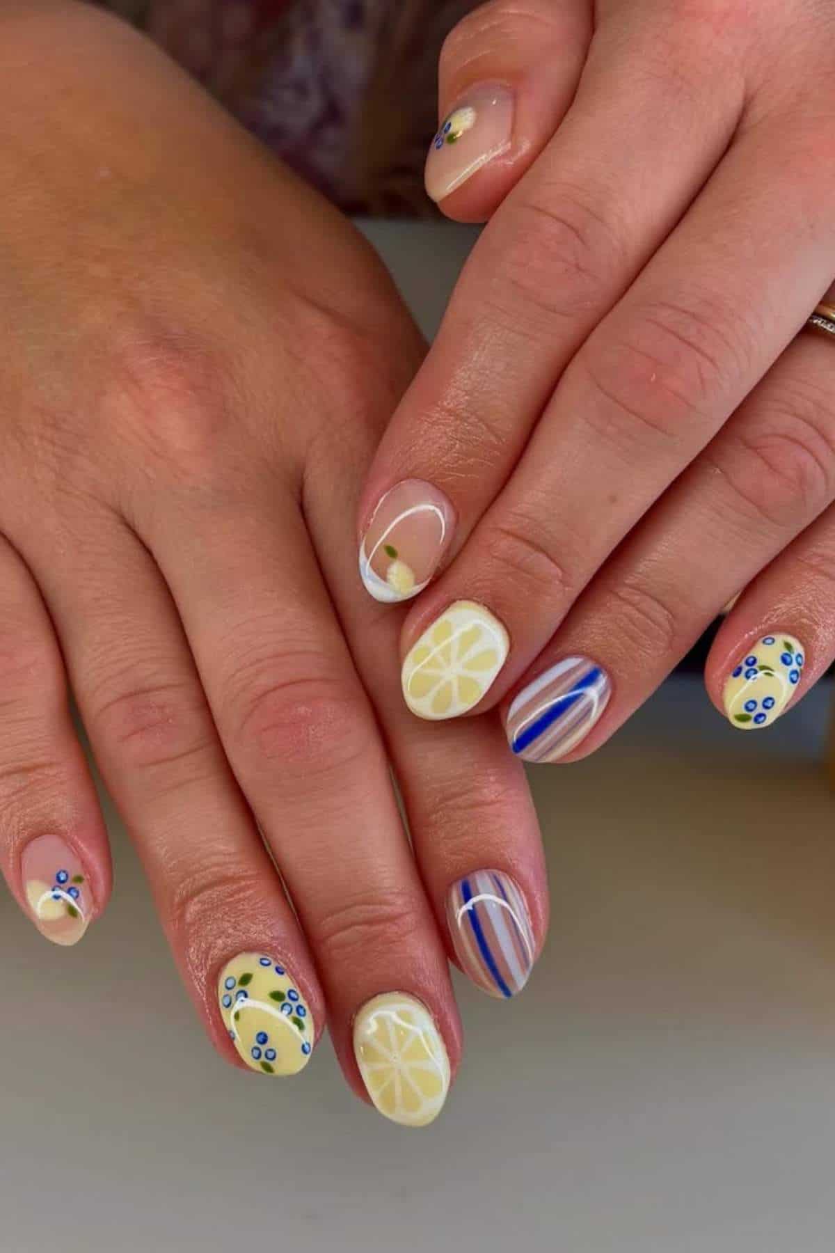 Hands with manicured nails featuring lemon slices, blue and white stripes, and small blue floral designs on a pale yellow and nude background.