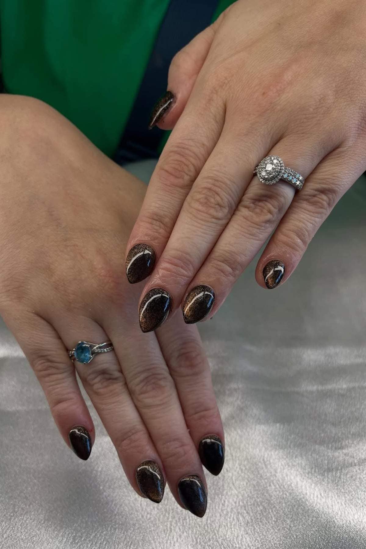 Two hands with almond-shaped, dark metallic nails and silver rings, one with a blue stone and the other with a clear stone, shown against a silver surface.