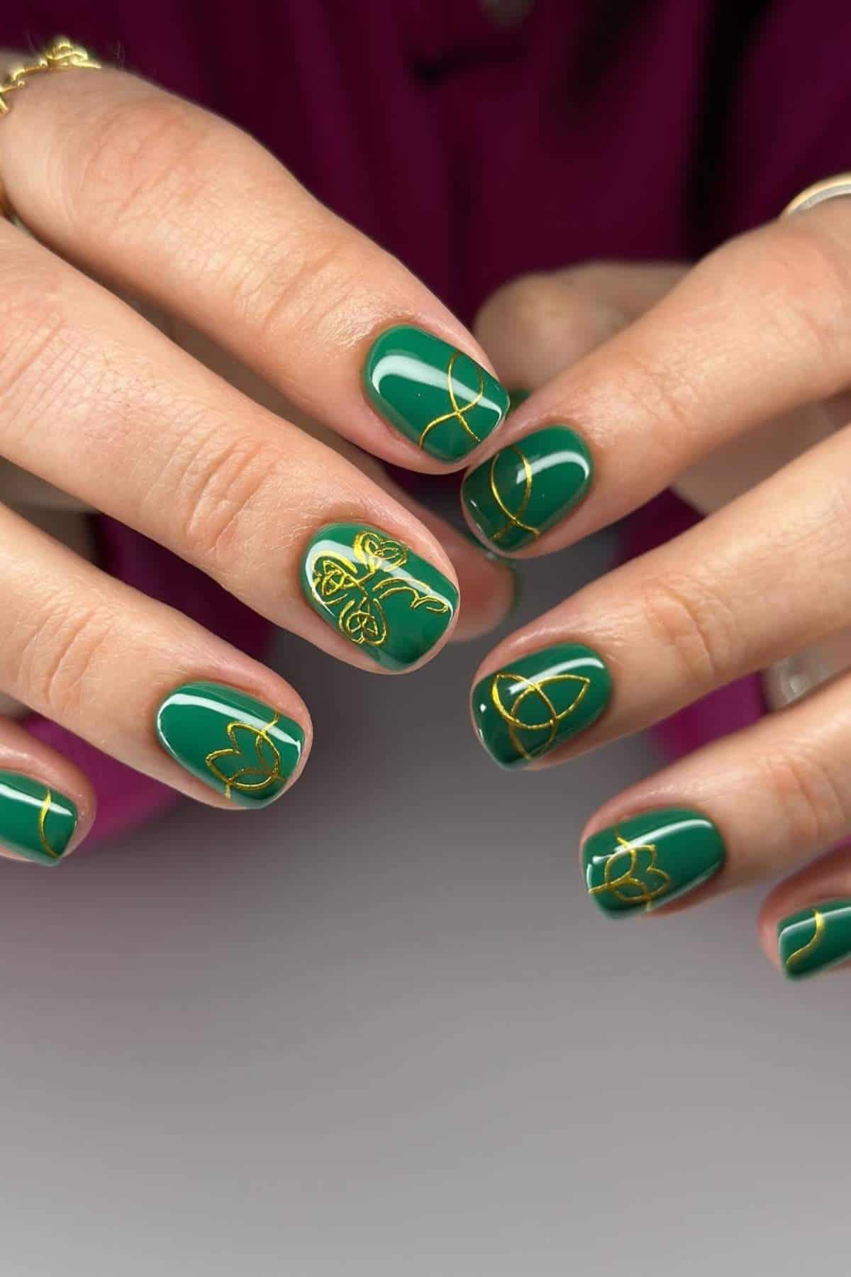 Hands with short, green-painted nails featuring delicate gold line designs and floral patterns against a neutral background.