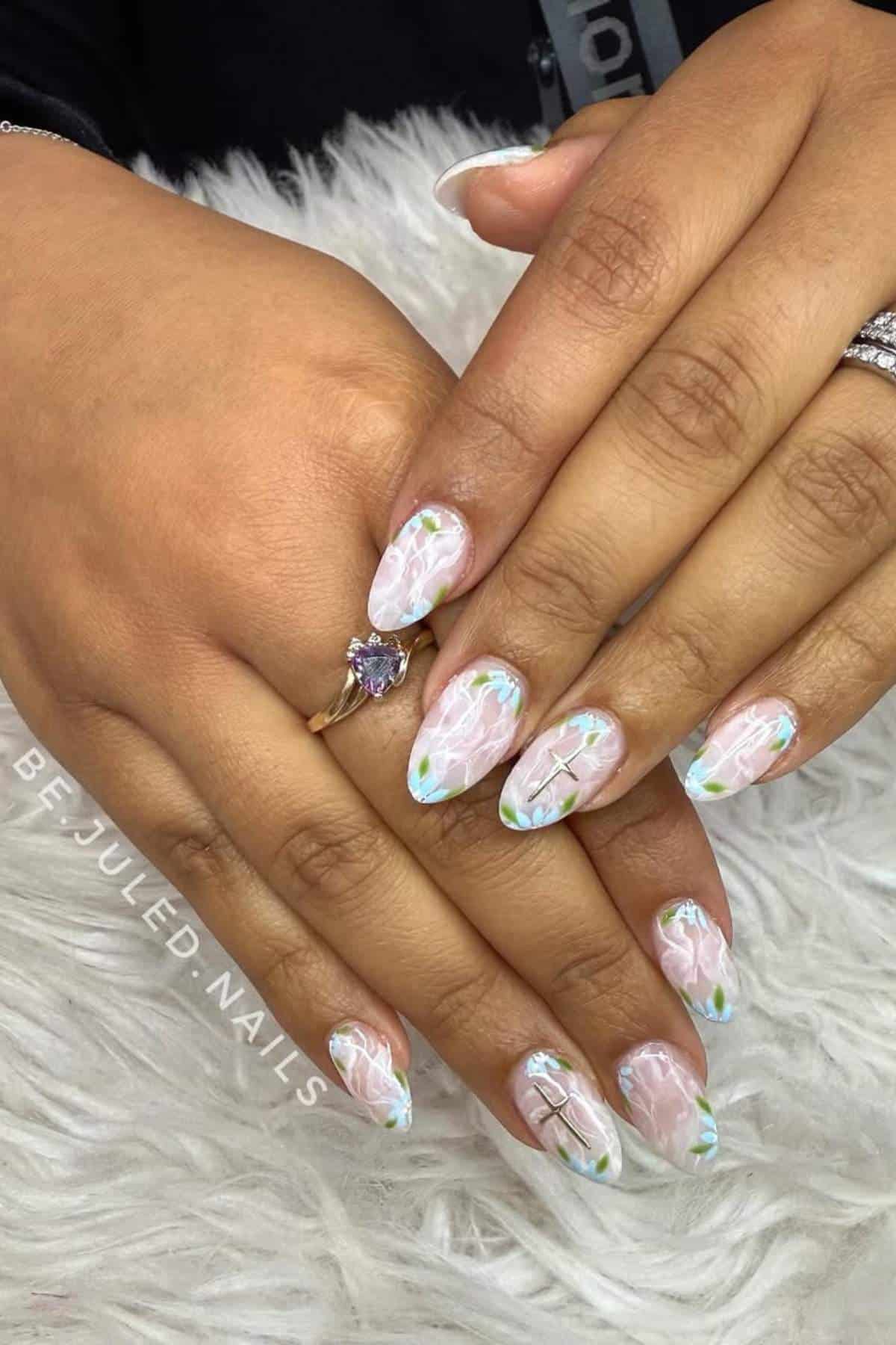 Hands with almond-shaped nails featuring a pale pink base and white and green floral designs, resting on a white furry surface. A ring with a heart-shaped stone is worn on one finger.
