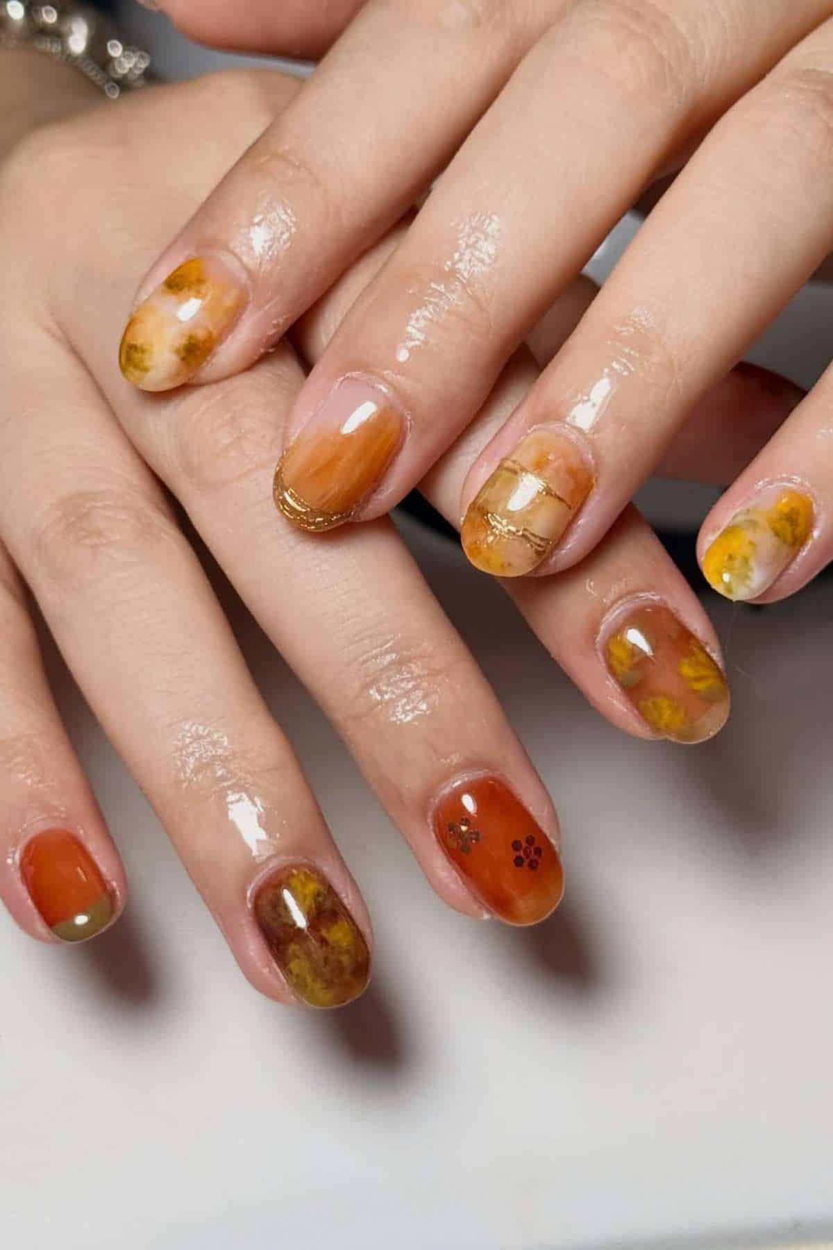 Close-up of hands with glossy nail art featuring warm orange, gold, and brown tones, marbled patterns, and subtle decorative accents on each fingernail.
