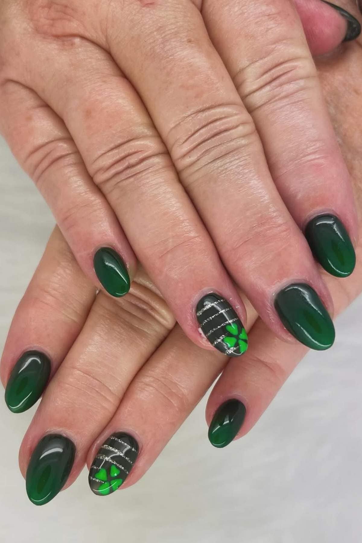 Hands with almond-shaped nails painted green, featuring accent nails with silver stripes and shamrock clover designs.
