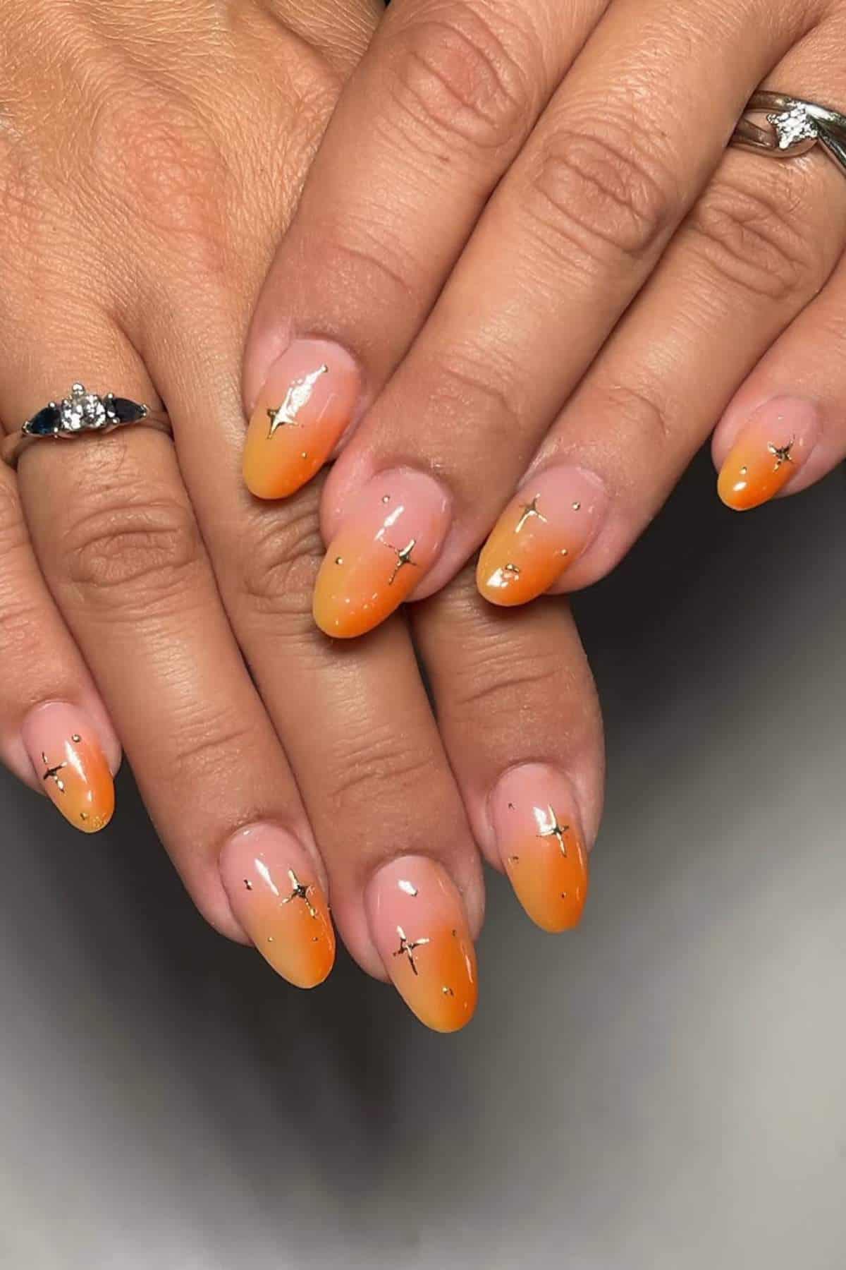 Hands with almond-shaped nails featuring orange ombre polish and small gold foil accents. Two rings are visible on the fingers.