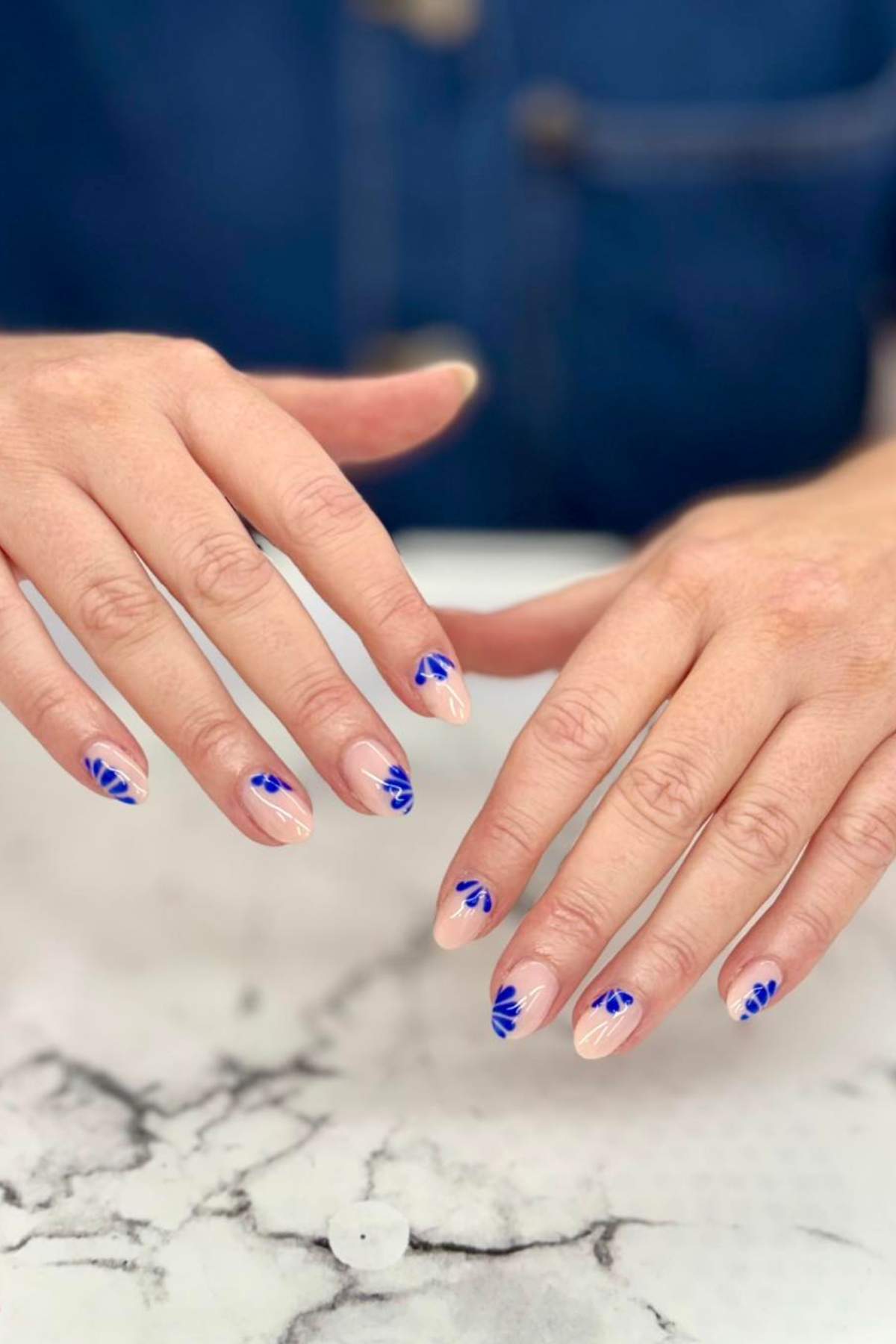 Two hands with almond-shaped nails featuring a nude base and blue leaf-like designs on a marble surface.