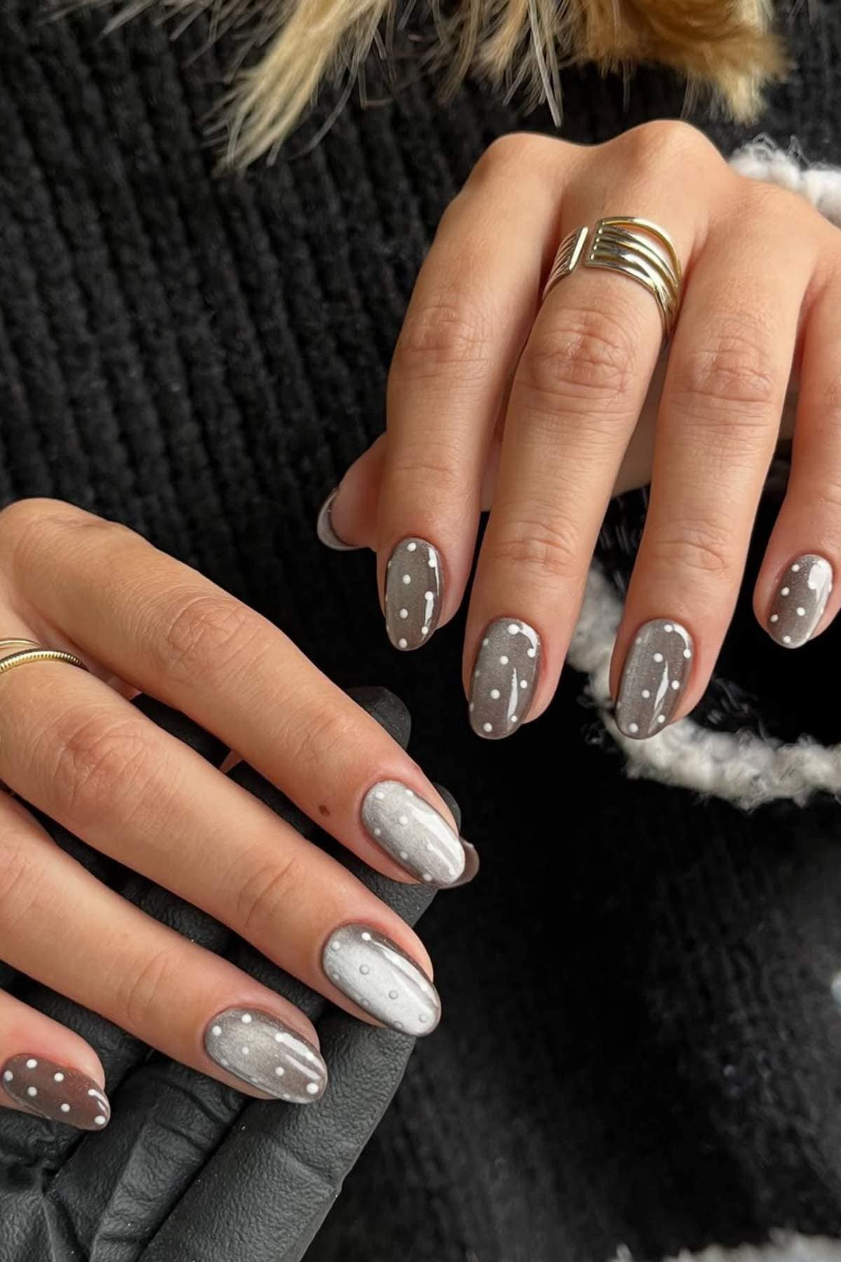 Hands with medium-length almond-shaped nails painted in shades of gray and white, featuring polka dot and line designs. Person wears gold rings and a black textured sweater.