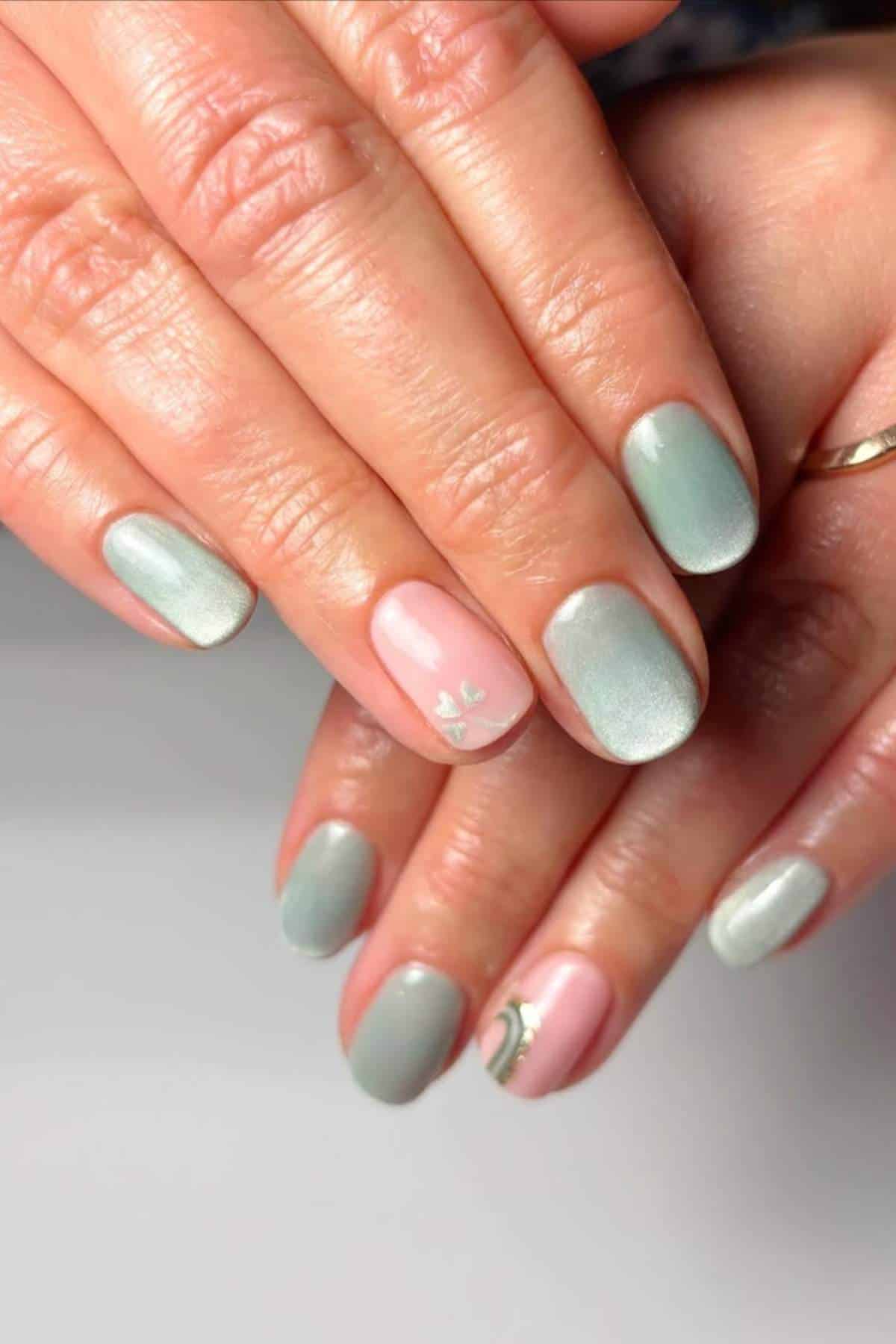 Hands with short, oval-shaped nails painted in light green and pink polish; two nails feature small, delicate white floral designs.