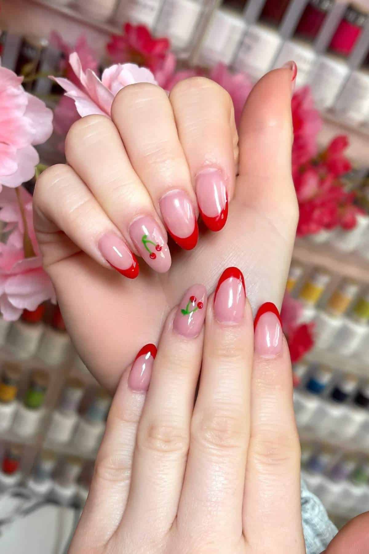 Hands with almond-shaped nails featuring red French tips and small cherry designs, held against a background with pink flowers and colorful nail polish bottles.