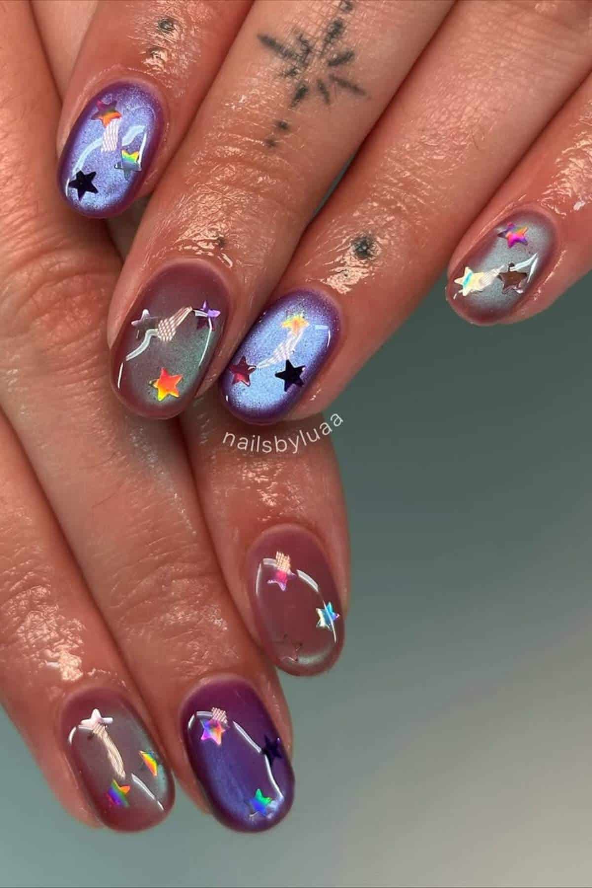 Hands with iridescent purple nails featuring star and ribbon designs, small finger tattoos, and a large star tattoo on one hand.