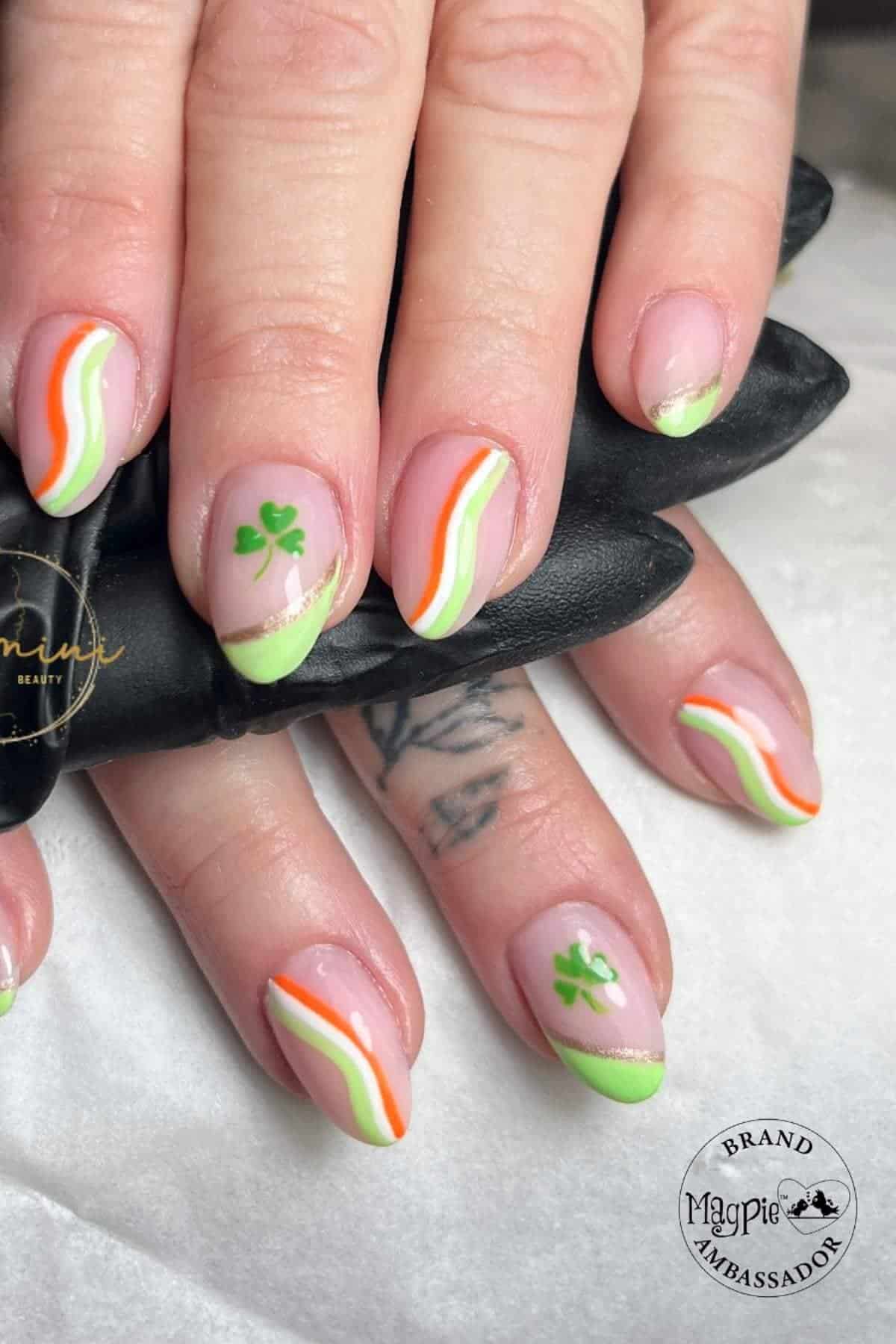 Hands with light pink nails featuring green, orange, and white wavy tips and small green shamrock designs on some nails.