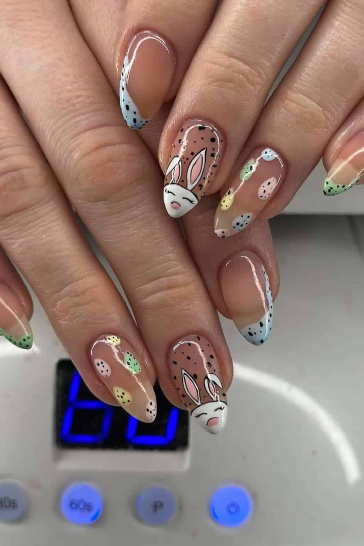 Close-up of hands with long, almond-shaped nails featuring pastel French tips, polka dot designs, and a white bunny face painted on two accent nails.