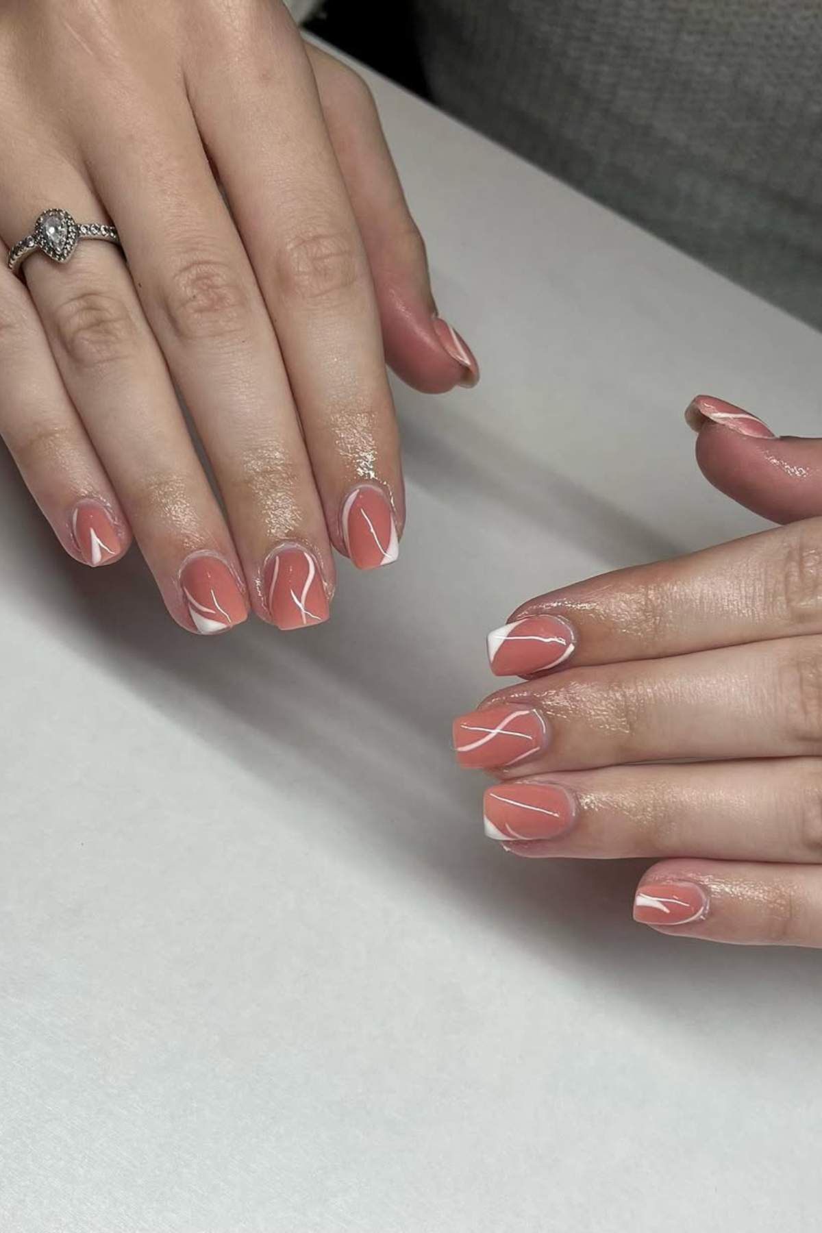 Hands with short, square-shaped nails painted in a peach-pink color with white abstract line designs and white tips, resting on a white surface.