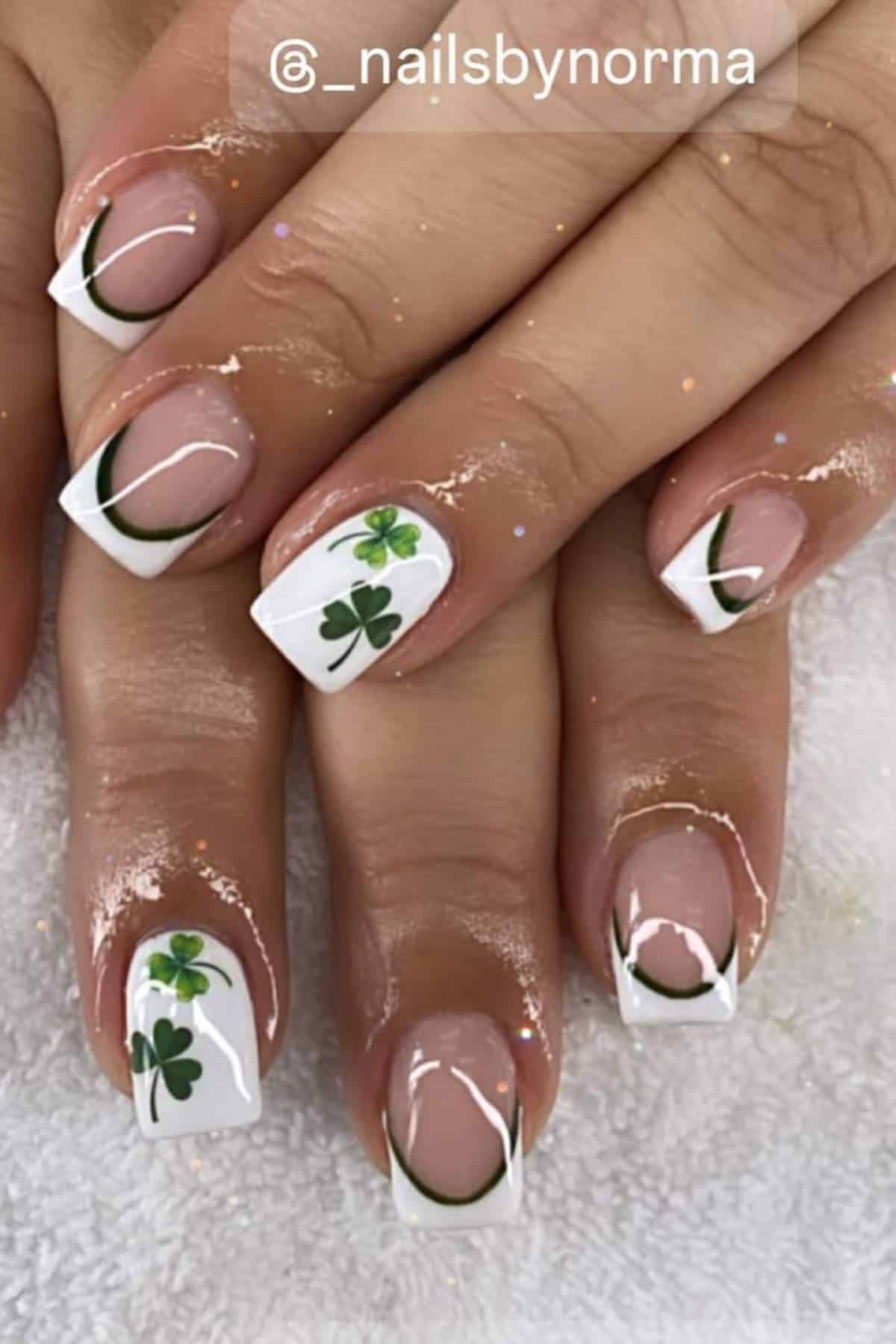 Close-up of hands with manicured nails featuring white French tips, green clover leaf designs, and green curved lines on a white towel background.