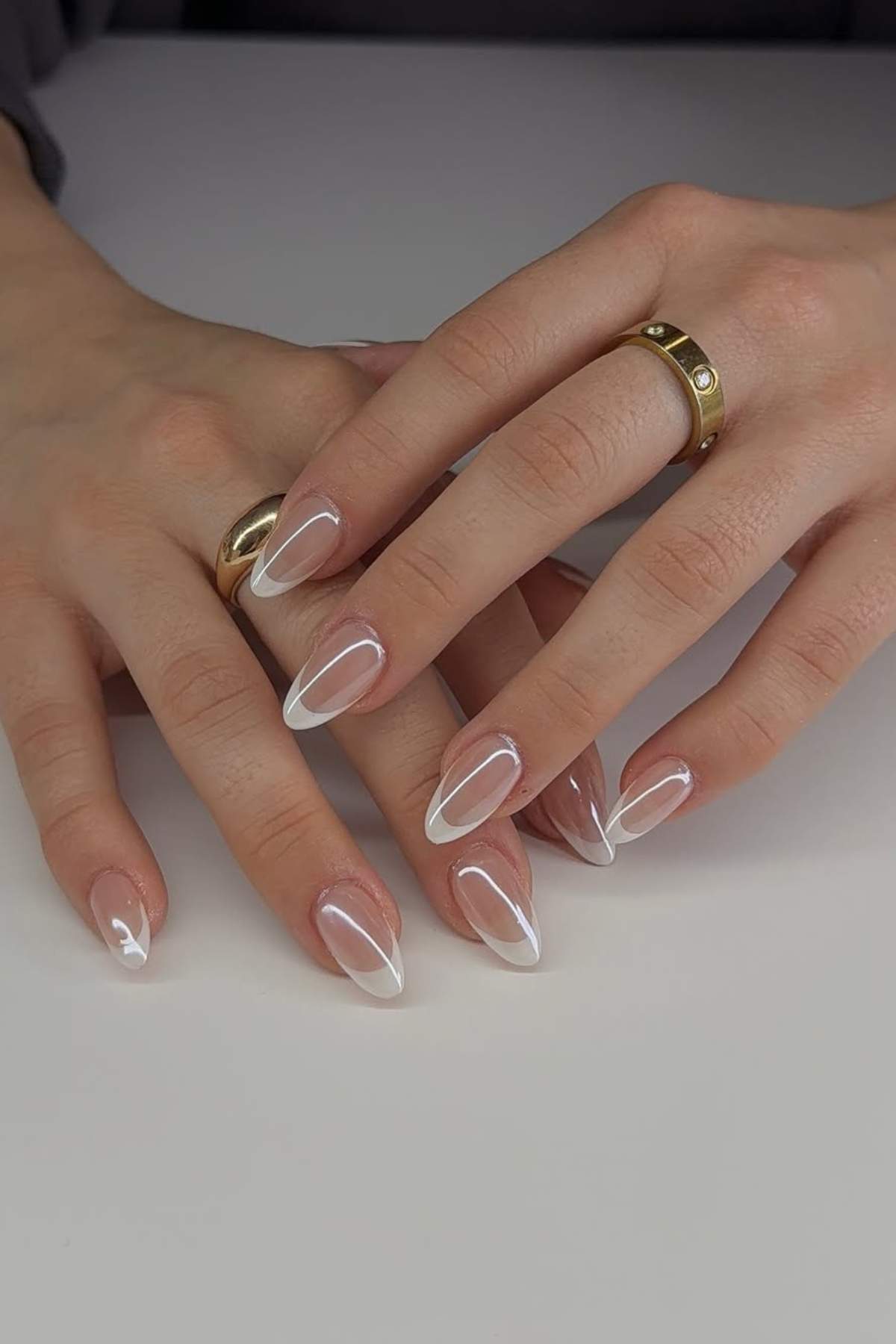 Hands with almond-shaped nails featuring a white French tip design rest on a white surface; gold rings are worn on both hands.