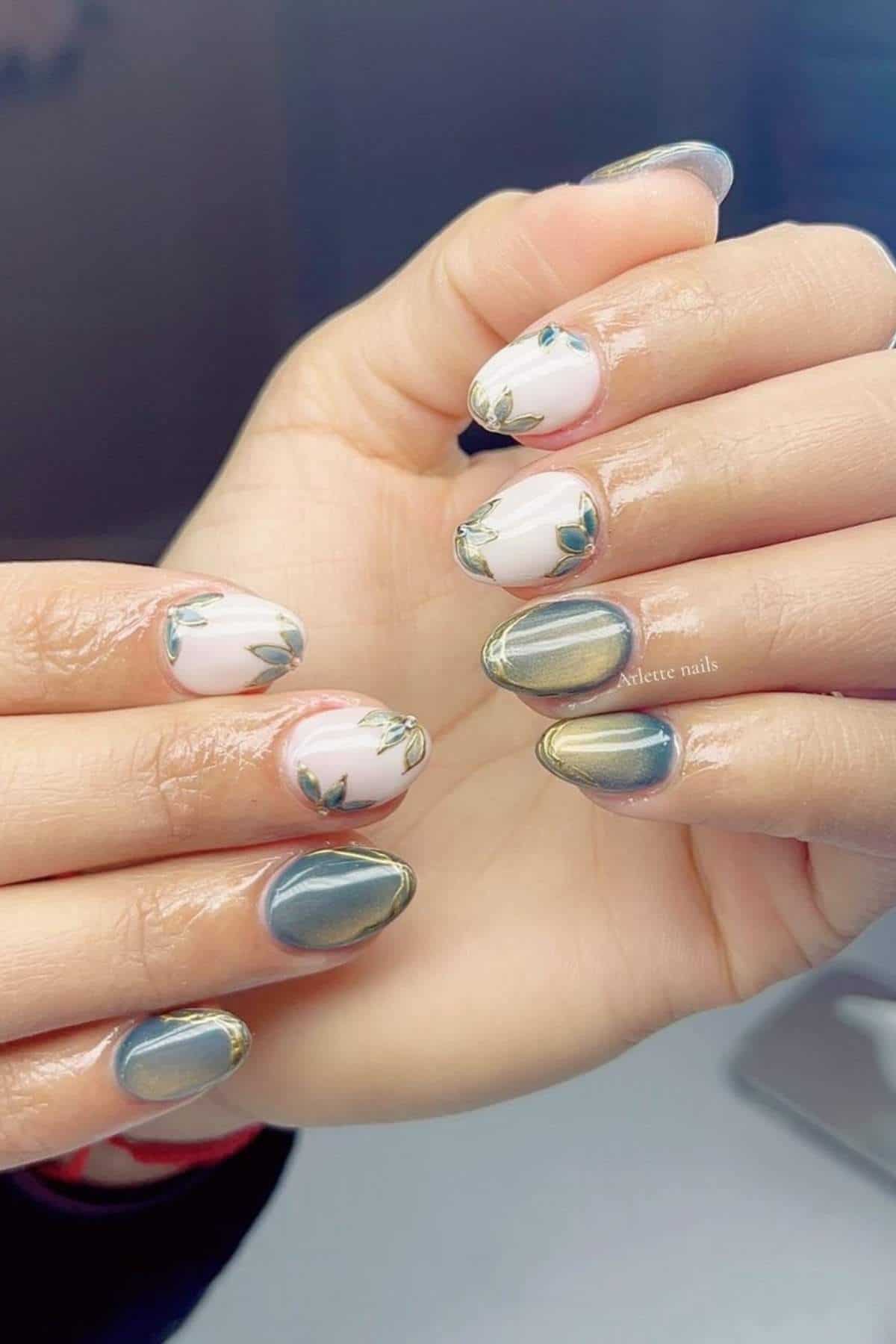 Hands with manicured nails featuring a metallic green polish with gold tips on some nails, and a white base with green leaf designs on others.