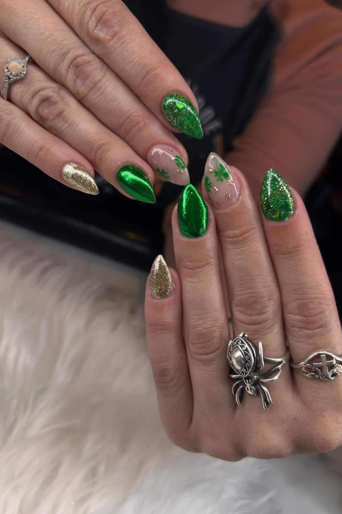 A hand with almond-shaped nails featuring green glitter, gold glitter, and clear nails with green shamrock designs. The person wears silver rings and a diamond ring.