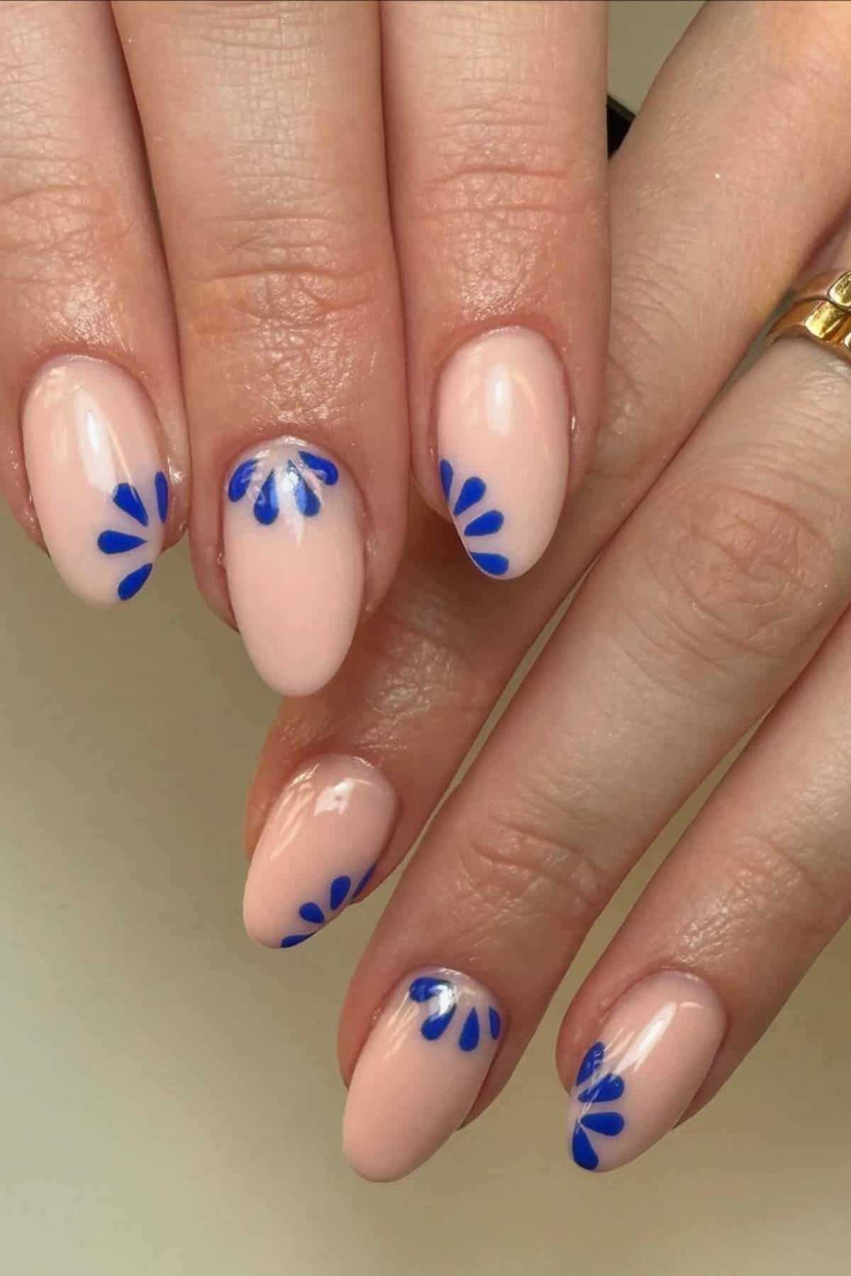 Hands with almond-shaped nails painted light pink, featuring blue petal-like designs near the cuticles on each nail.