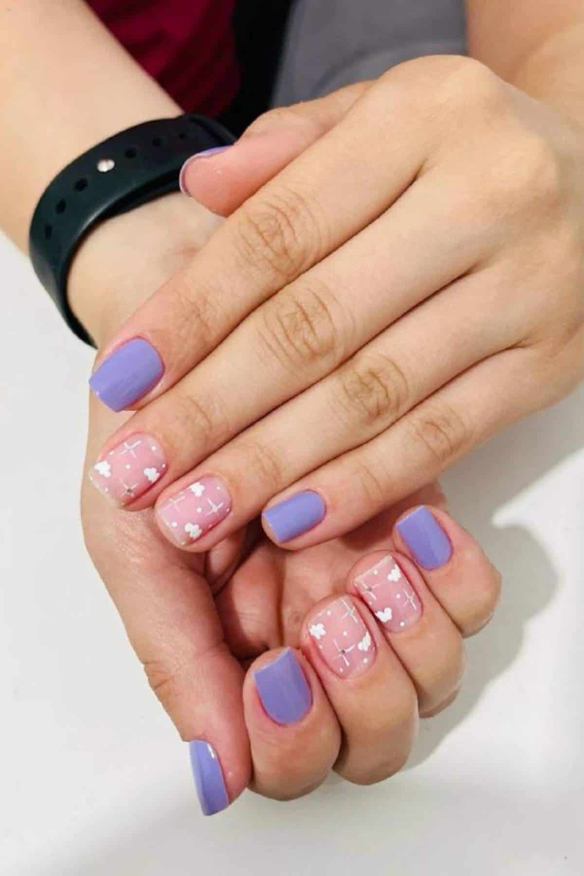 Close-up of hands with short, square-shaped nails painted in lavender and clear polish with white heart designs on two accent nails. Person is wearing a black wristband.