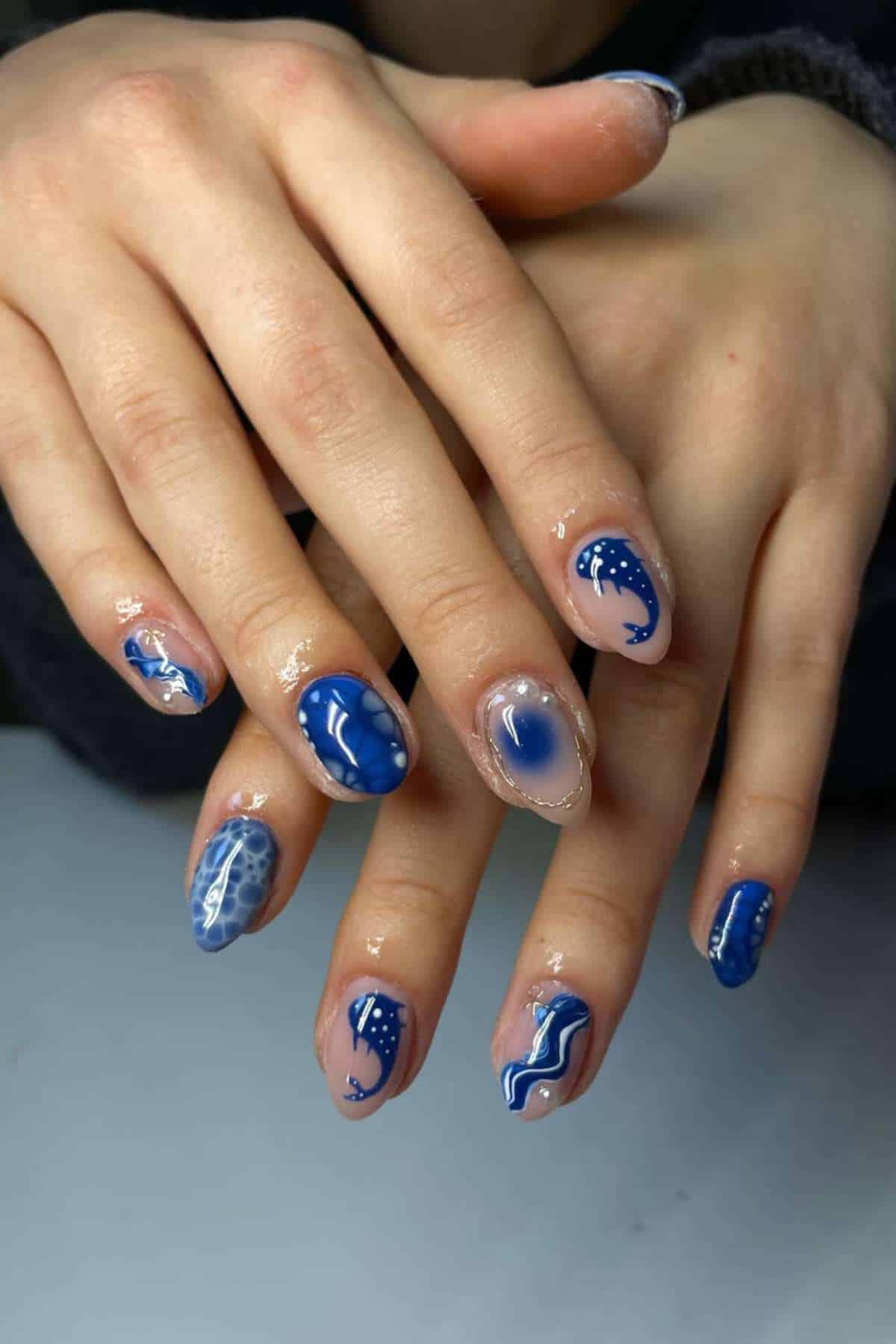 Hands with almond-shaped nails featuring blue and white ocean-themed nail art, including waves, whales, and abstract patterns.