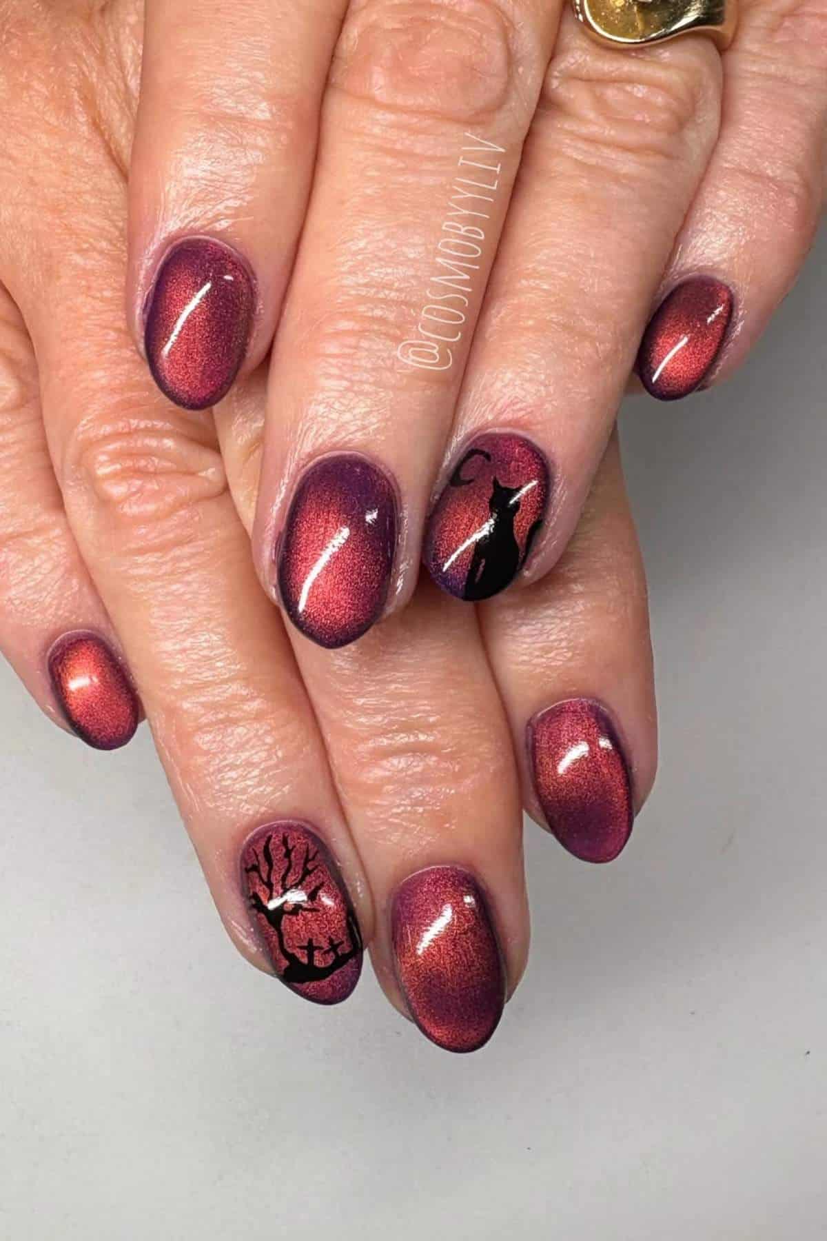 Hands with deep red, shiny ombre nails featuring black designs of a cat on one nail and a tree with branches on another.