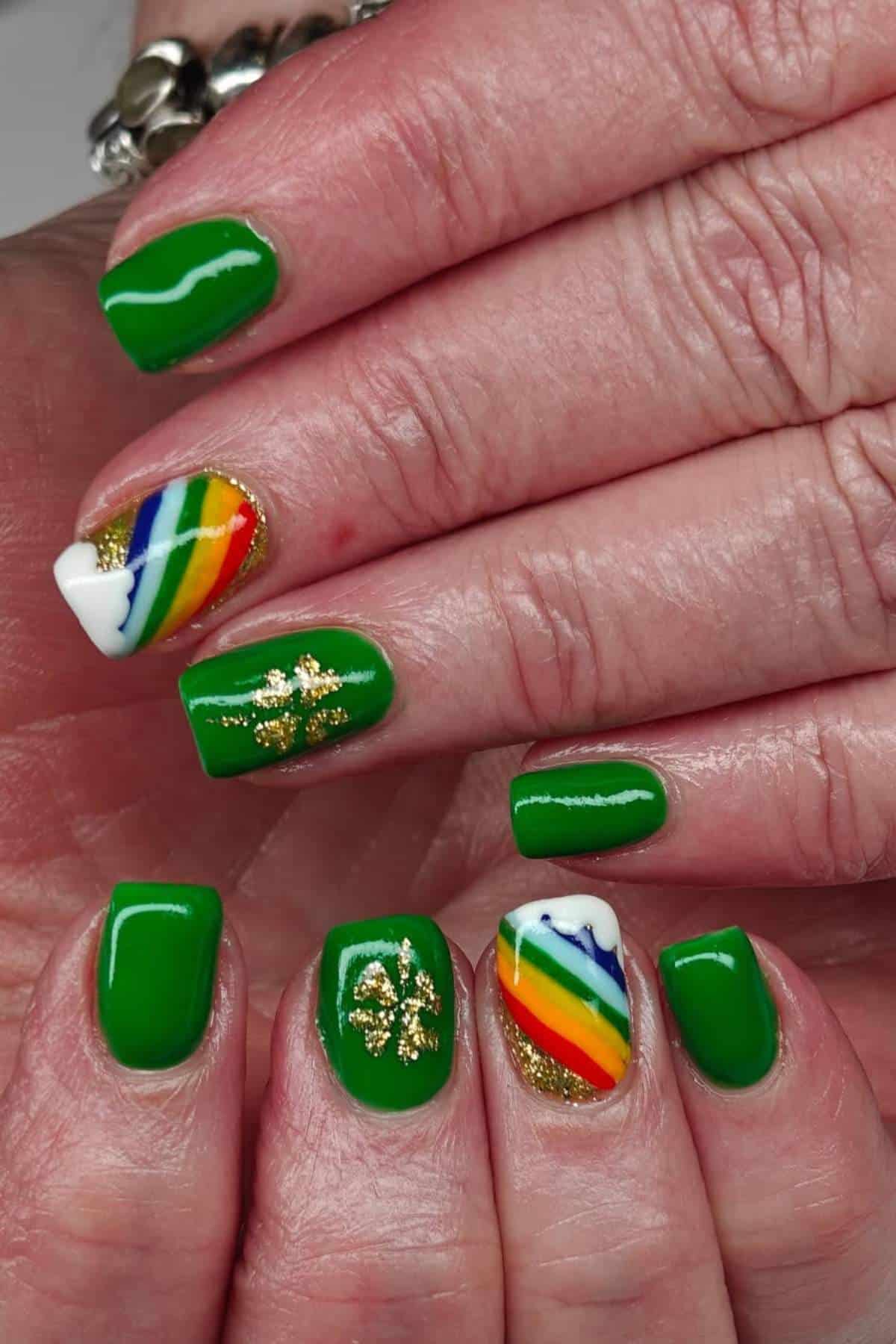 Hands with short, square-shaped nails painted green; some nails feature gold shamrock designs and others have rainbow patterns with white tips and gold glitter accents.