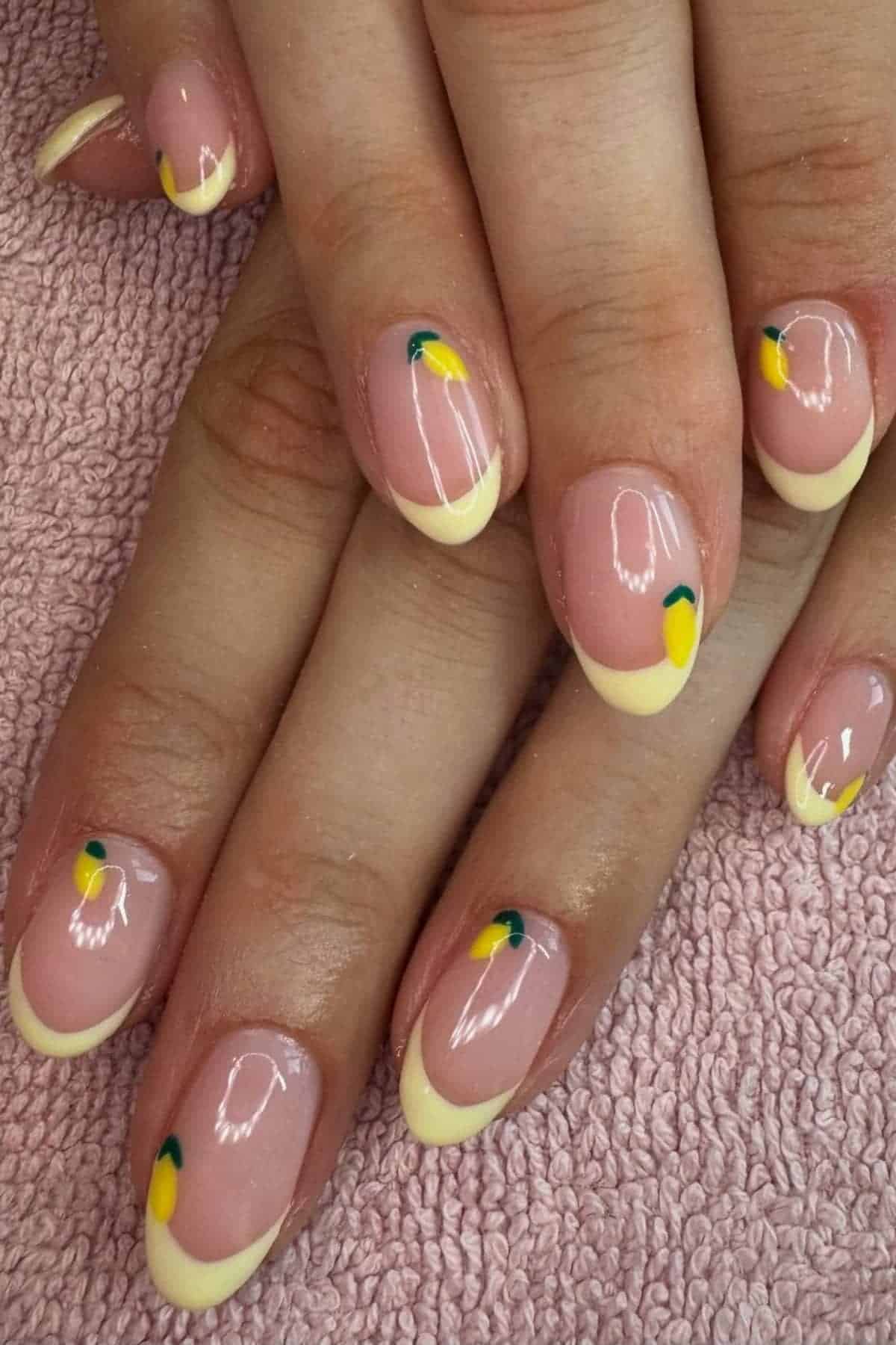 Hands with manicured nails featuring a pale yellow French tip and small lemon slice designs, resting on a pink textured towel.