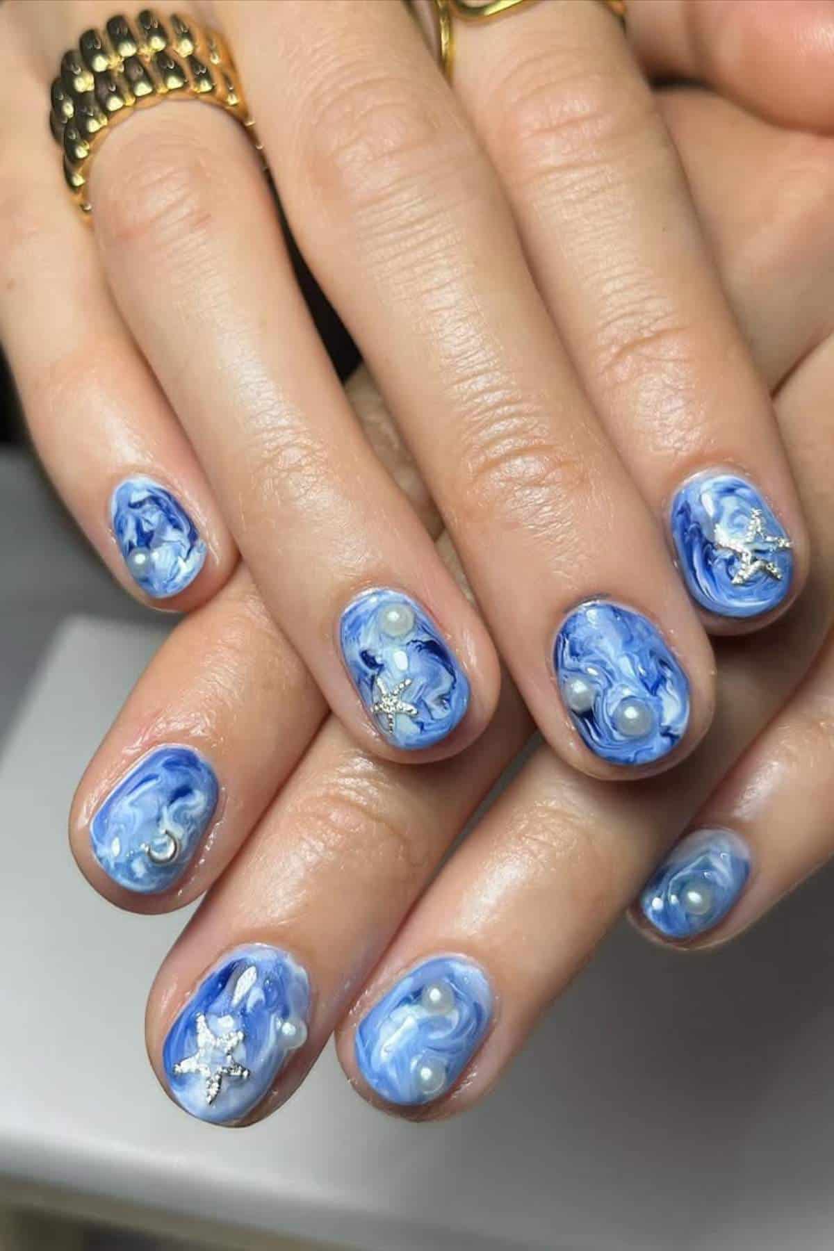 Close-up of hands with short, oval nails painted in blue and white marble design, decorated with small pearls and silver star embellishments.
