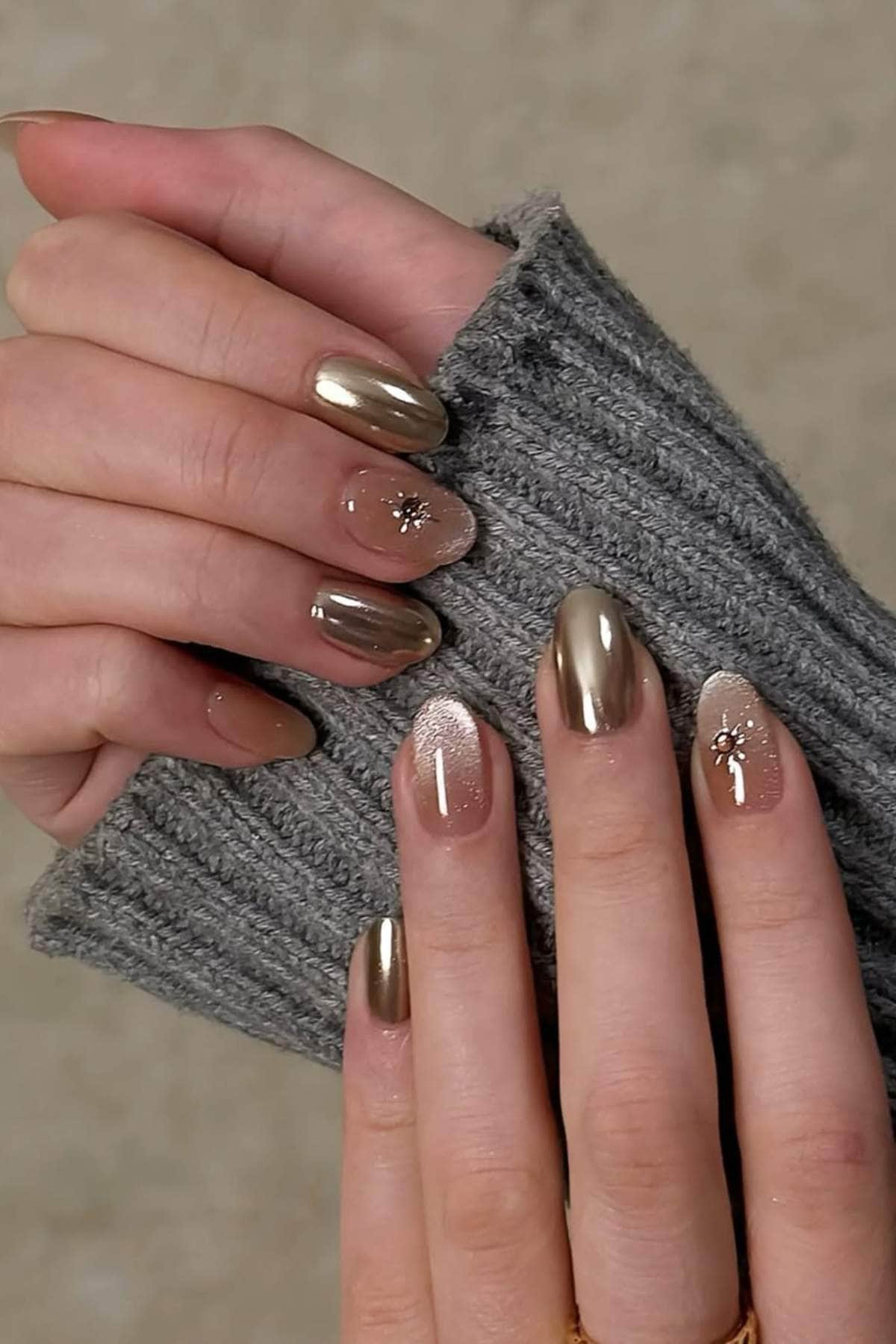 Close-up of hands with gold metallic and nude nails featuring small rhinestone and glitter accents, partially covered by a gray ribbed sleeve.