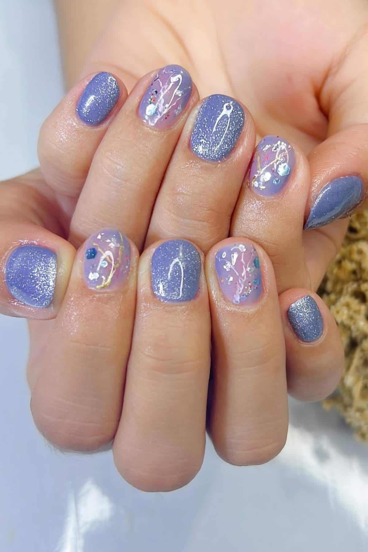 Close-up of hands with short, rounded nails painted in purple and glitter polish, some with abstract designs, sparkles, and small decorative accents.