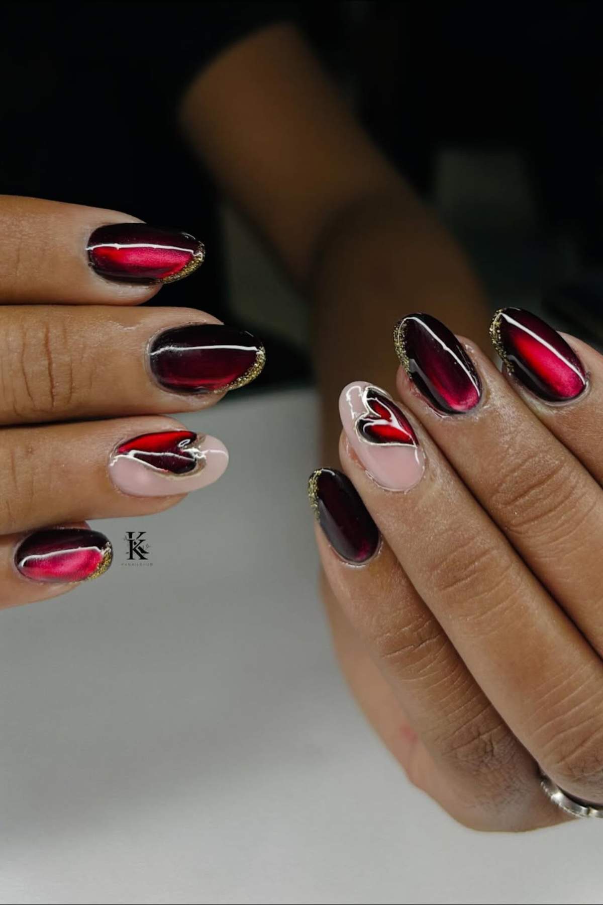 Hands with almond-shaped nails featuring glossy red and black ombre polish, gold accents, and heart designs on two accent nails.