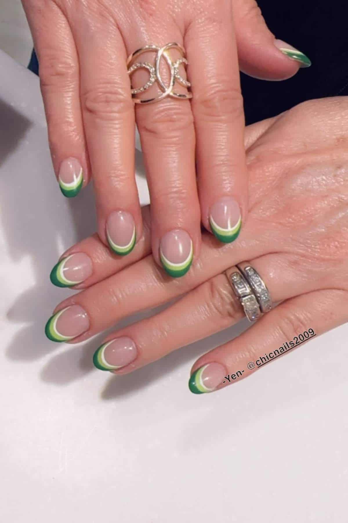 Hands with nude nails featuring green and white French tips, adorned with silver and gold rings.