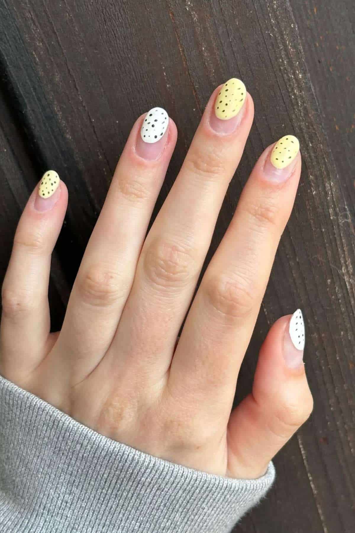 A hand with short, almond-shaped nails painted in alternating yellow and white polish, each with small black polka dots, against a wooden background.