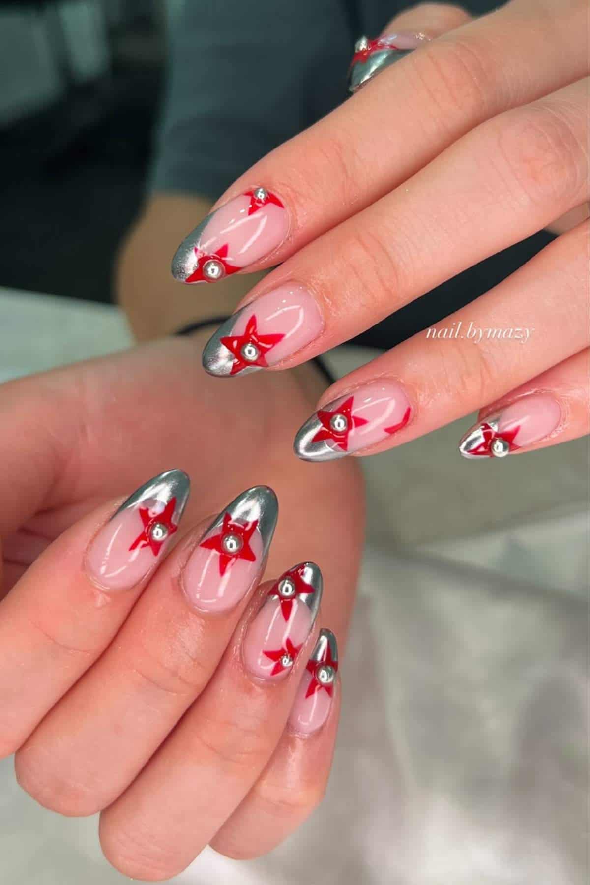 Hands with almond-shaped nails, featuring silver chrome tips and red star designs with small rhinestone accents on a light pink base.