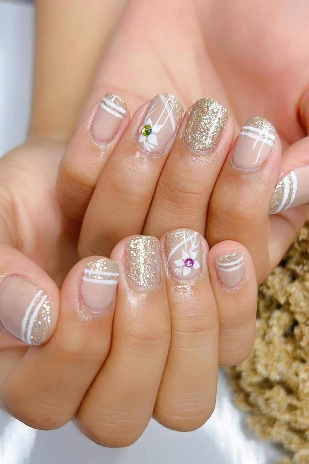 Hands with short nails featuring gold glitter polish, white line designs, and small rhinestone accents on a clear or nude base.