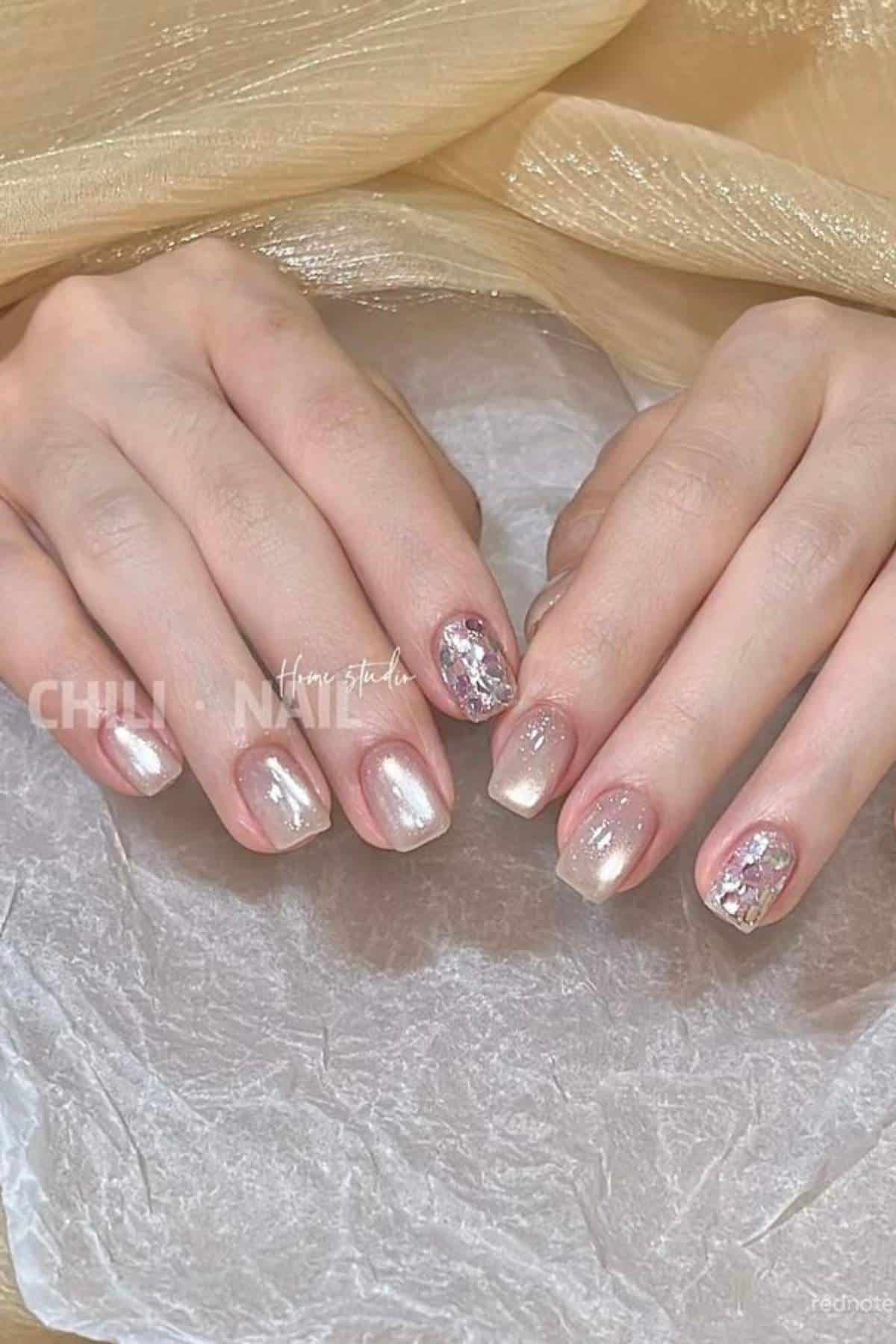 A pair of hands with short, square-shaped nails painted in pearlescent pink and white shades, featuring accent nails with glitter and foil decorations.