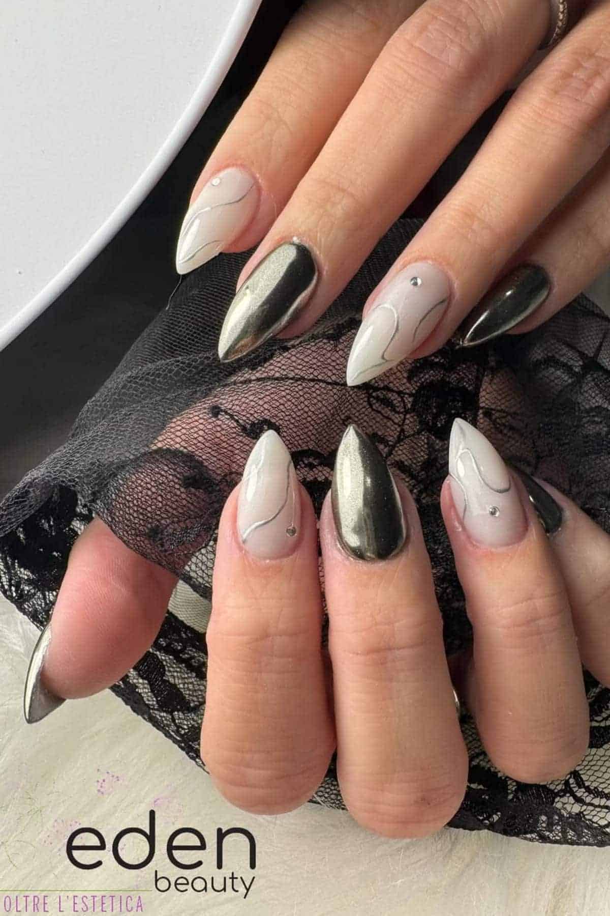 Close-up of hands with almond-shaped nails featuring a black chrome and nude design with abstract lines and small rhinestone accents.