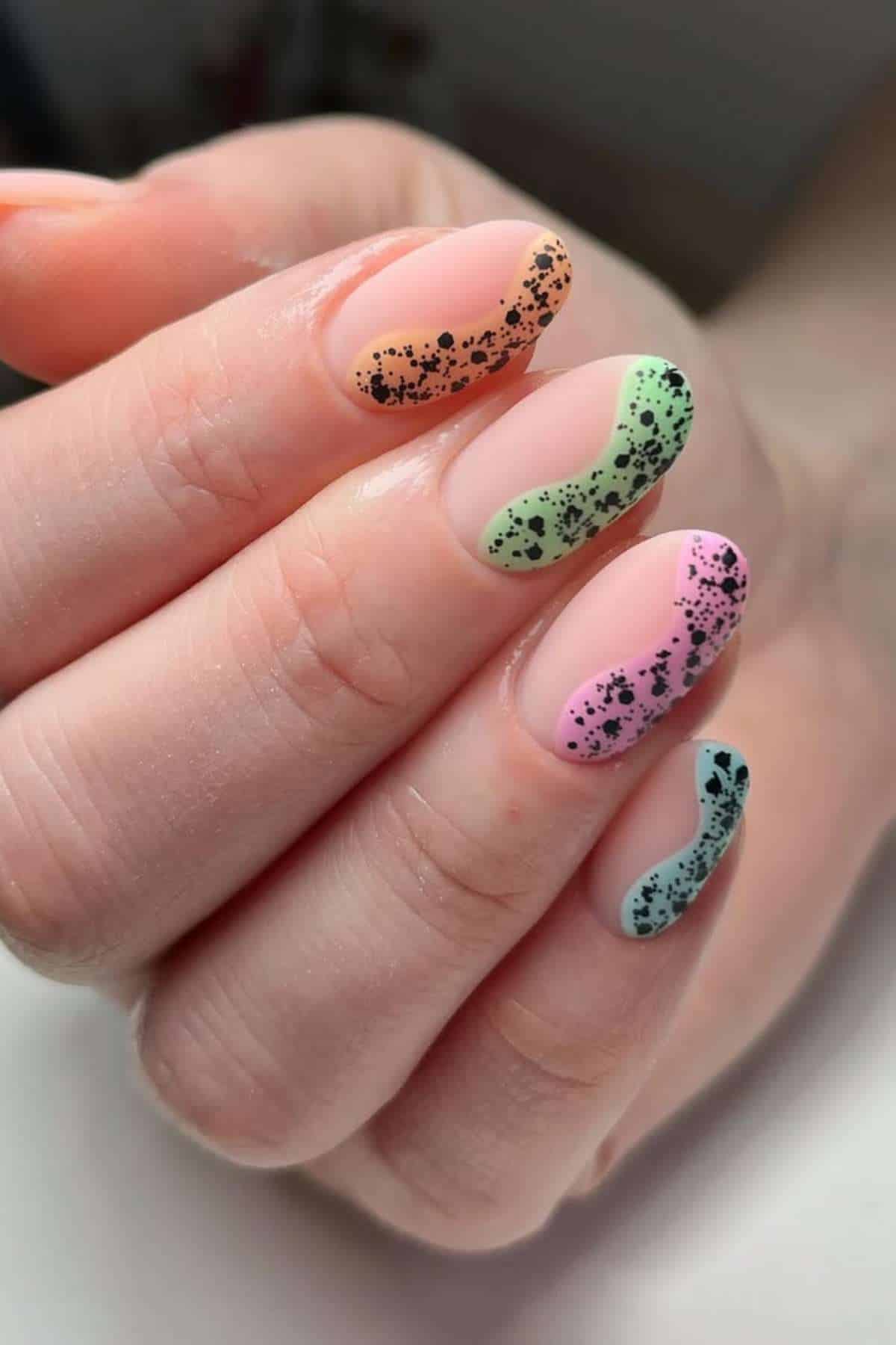 A hand with long oval nails, each painted a different pastel color with a black speckled wavy line design on the tips.