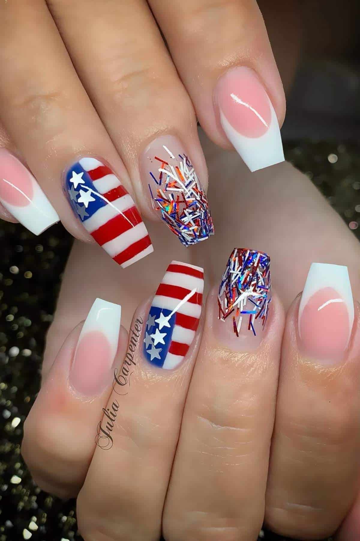 A hand with long square French tip nails, two nails featuring an American flag design, and two with red, white, and blue glitter accents.