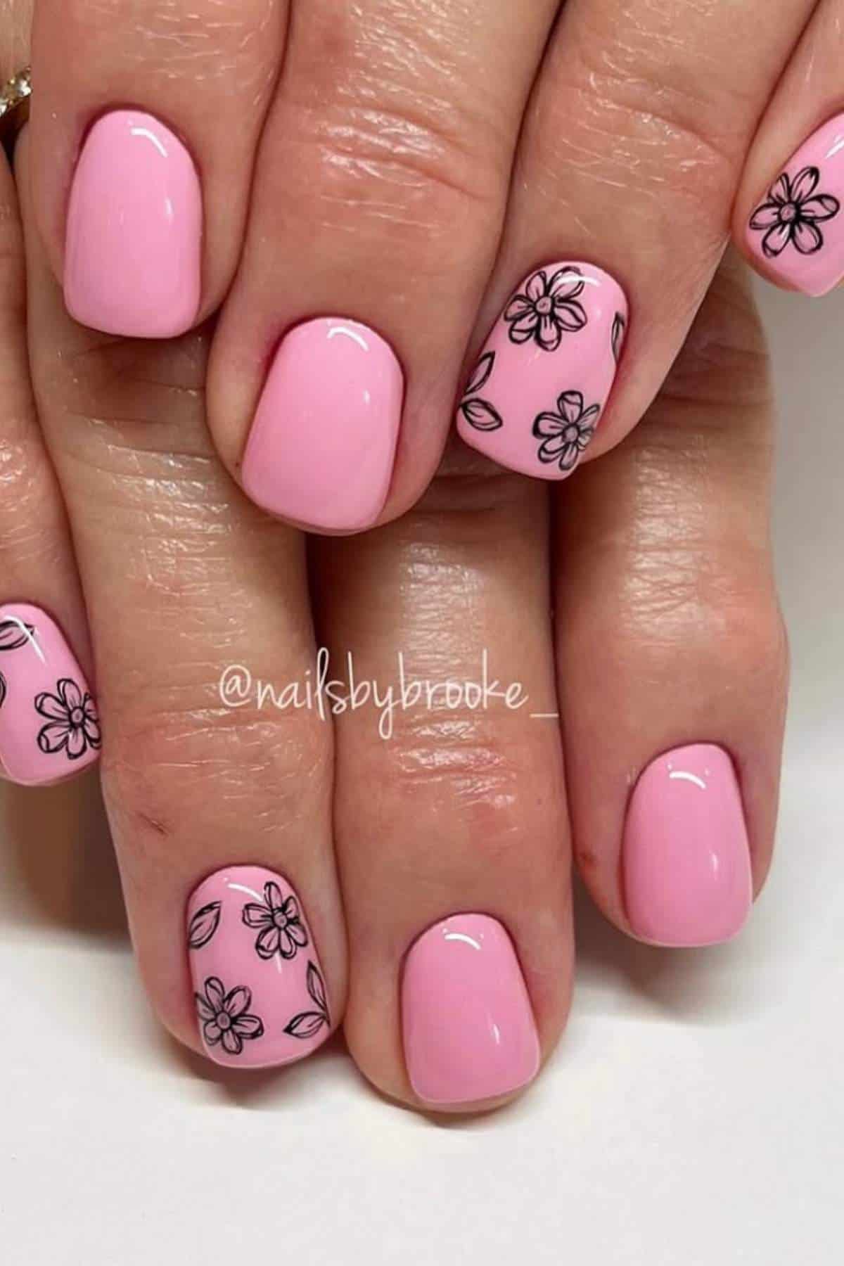 Hands with short, square-shaped nails painted light pink; some nails feature black outlined flower designs.