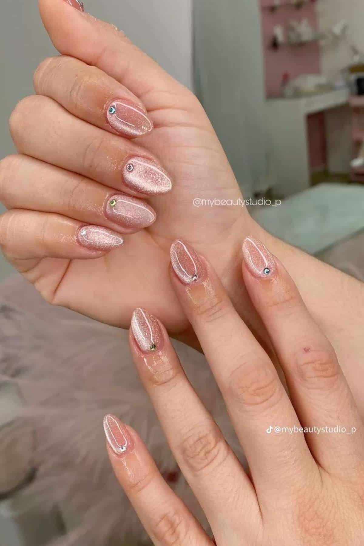 Hands with almond-shaped, nude-pink nails featuring a subtle shimmer and small rhinestone accents near the cuticles.