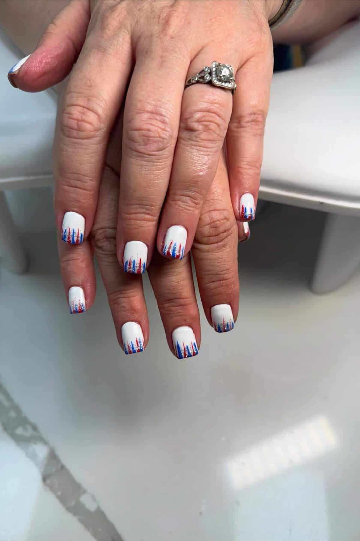 Two hands display white manicured nails with red and blue vertical line designs at the tips. The person wears a ring with a large stone on one finger.